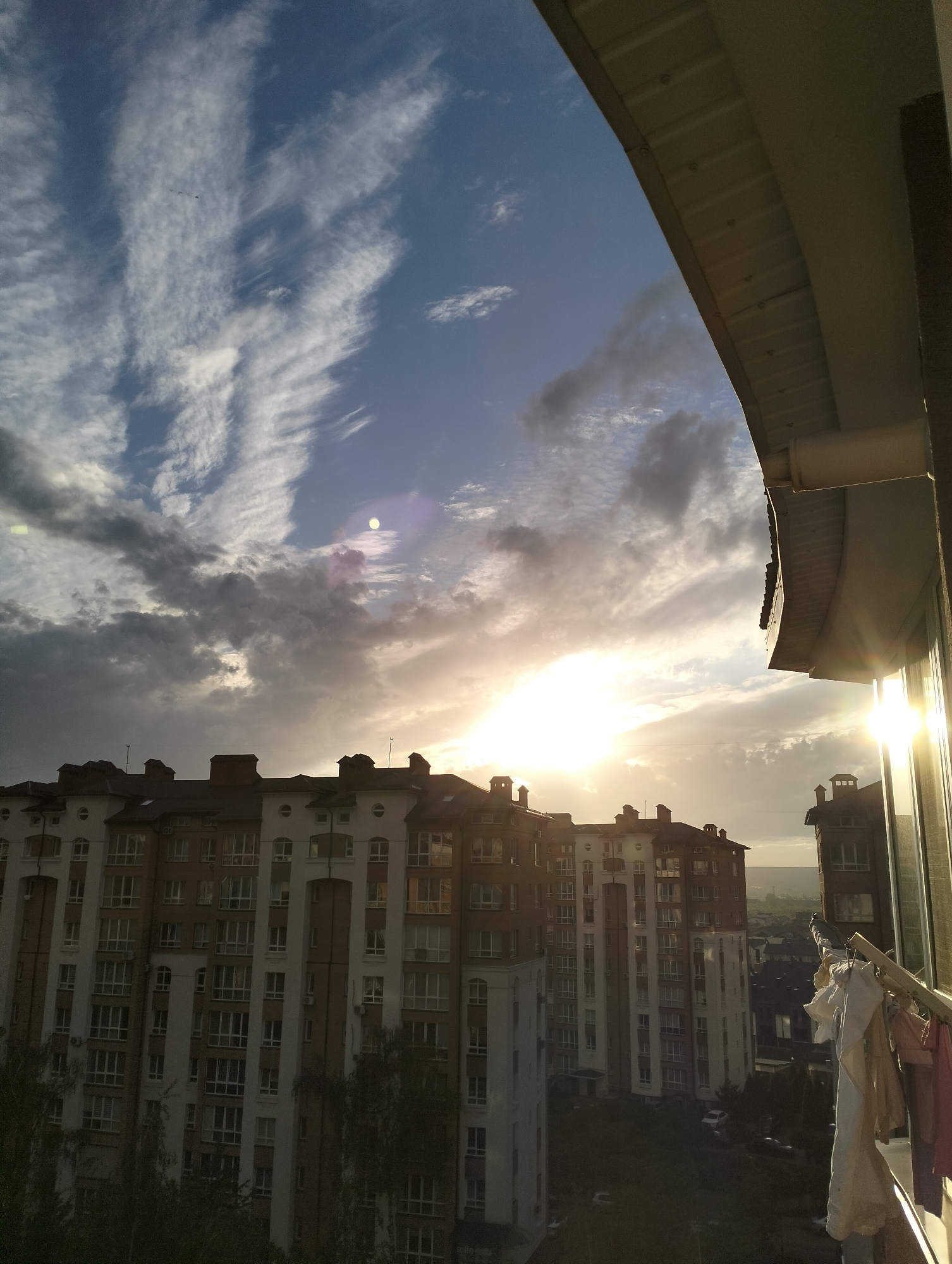 The image depicts a cityscape in Krykhivtsi, Ukraine, viewed from a high vantage point, likely a balcony or high-rise apartment. The foreground includes a building with several stories and a combination of red brick and white facade. A clothesline with laundry hangs on the right side of the frame, attached to a balcony railing. Sunlight streams through a window, creating a lens flare.
The sky is filled with a mix of clouds, some wispy and others dense and gray. The sun is obscured but visible, casting a bright glow. Further in the background, there are more buildings and possibly distant hills or mountains. The overall environment suggests a residential area on the outskirts of a city, with a combination of urban and natural elements. No people are visible in the image.