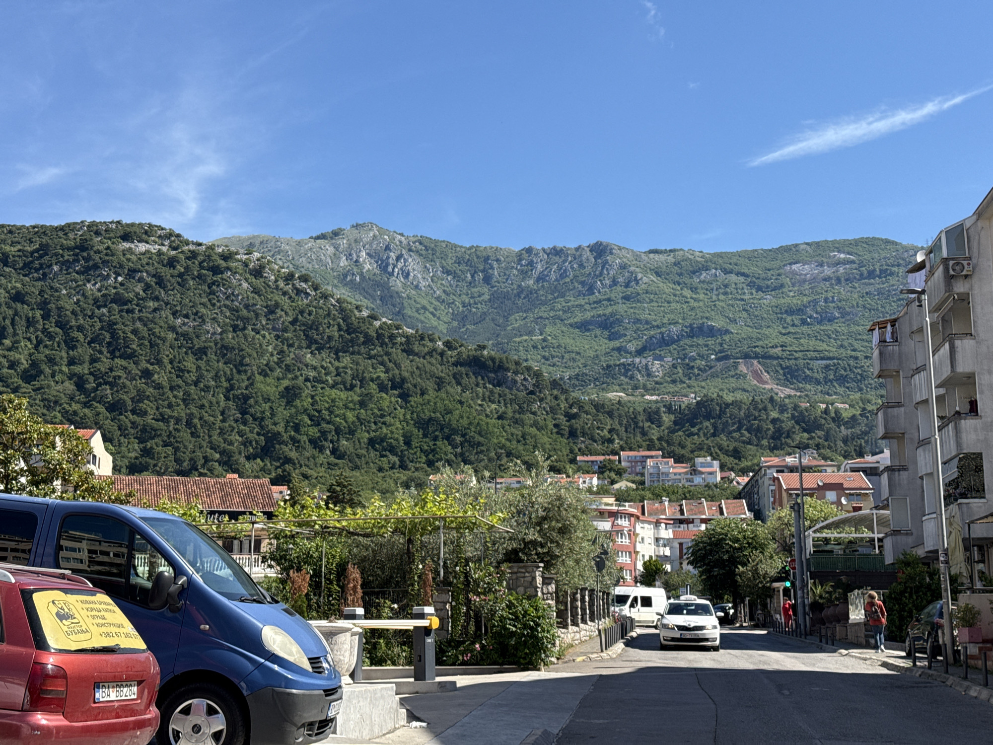 The image depicts a street scene in Budva, Montenegro on a clear, sunny day. The focus is a street leading towards a distant mountain range densely covered in green vegetation. The sky is a vivid blue with sparse wisps of clouds.
In the foreground, to the left, are several parked cars: a red hatchback with license plate "BA BB284" and a blue minivan. A sticker on the back window of the red car advertises metalwork services, including fabrication of fences. Adjacent to the cars, a low stone wall with a gated entrance indicates a private property.
The street itself is empty except for a white taxi cab and a white van driving in the distance. On the right side of the road, a pedestrian is walking on the sidewalk towards the viewer, while another pedestrian in a red shirt walks further down the road. To the right side is a multistory building with balconies. In the background, houses with red tiled roofs are nestled against the mountain backdrop.