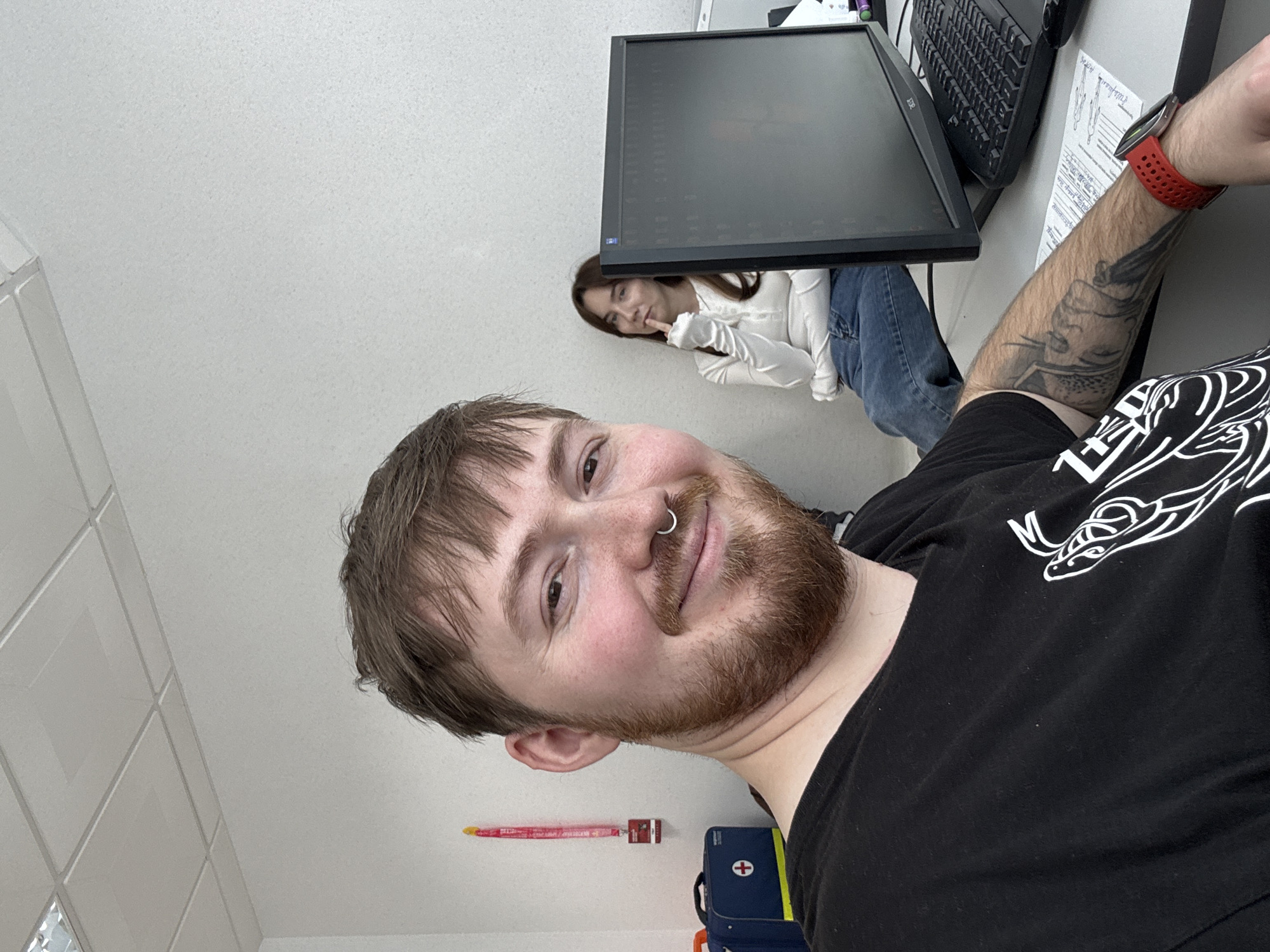 A person is holding the camera in a selfie-style shot. The person has light skin, brown hair and beard, a nose ring, and is wearing a black t-shirt. A tattoo is visible on the person's left forearm. The background includes a computer monitor, keyboard, and a woman with long brown hair wearing a white shirt and blue jeans. The background also shows a red thermometer and a blue first aid kit on a white wall with ceiling tiles. The location is possibly inside an office or workspace. Based on the metadata, the location is Прикамский, Russia.