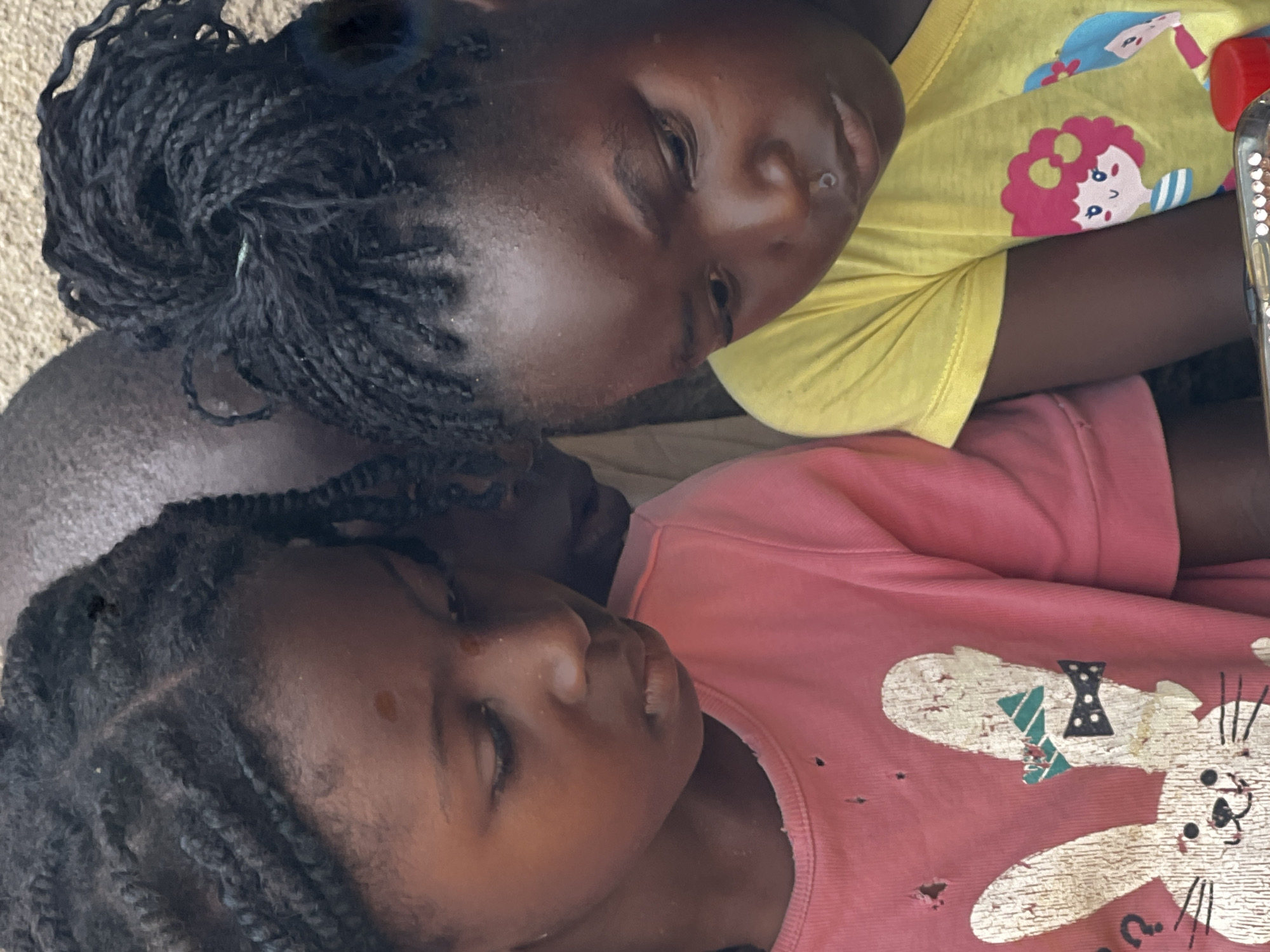 Two young girls are looking down at a device, likely a smartphone, in this close-up, outdoor shot. The girl on the left, wearing a pink t-shirt with a distressed bunny graphic, has her hair in braids and is positioned slightly behind and to the left of the other girl. The girl on the right, in a yellow t-shirt adorned with cartoon characters, has her hair in braids styled in a bun and is looking intently at the device. Both girls appear to be of African descent, with dark skin tones. The background is a textured wall, likely made of concrete or stucco. The lighting suggests it is daytime, with soft, diffused light. The overall atmosphere is one of focused engagement with the technology. The image is a tight crop, emphasizing the faces and upper bodies of the children. The scene depicts a common modern interaction, with technology playing a central role in the children's attention. The specific location in Jos, Nigeria is not ascertainable from the visual evidence alone, but the context suggests a typical urban or semi-urban setting.
