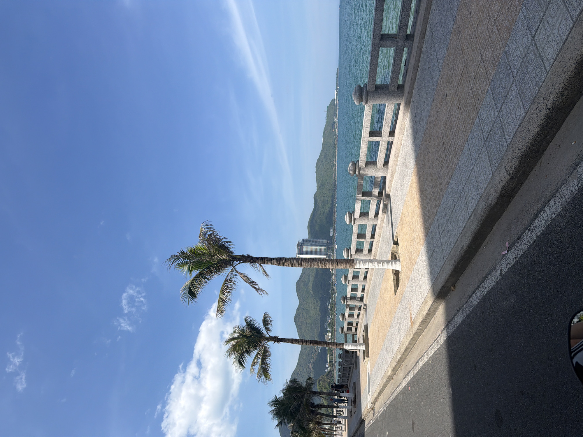 A wide shot captures a scenic coastal road in Nha Trang, Vietnam, under a bright blue sky dotted with sparse white clouds. The foreground features an asphalt road on the left, with a white dashed line marking the lane. A concrete curb separates the road from a paved pedestrian walkway. This walkway is tiled with alternating patterns of grey and tan squares and features a decorative concrete barrier with rounded posts overlooking the ocean. Several palm trees are planted along the walkway, their trunks wrapped in white material near the base.
Beyond the walkway, the turquoise ocean stretches to the horizon, meeting a range of verdant, tree-covered mountains in the distance. A tall, modern building with a light-colored facade is visible on the slopes of the mountains. A few boats are scattered on the water. A handful of people are visible on the walkway, appearing to be strolling or standing. The overall scene depicts a well-maintained public space by the sea, likely a popular tourist spot.