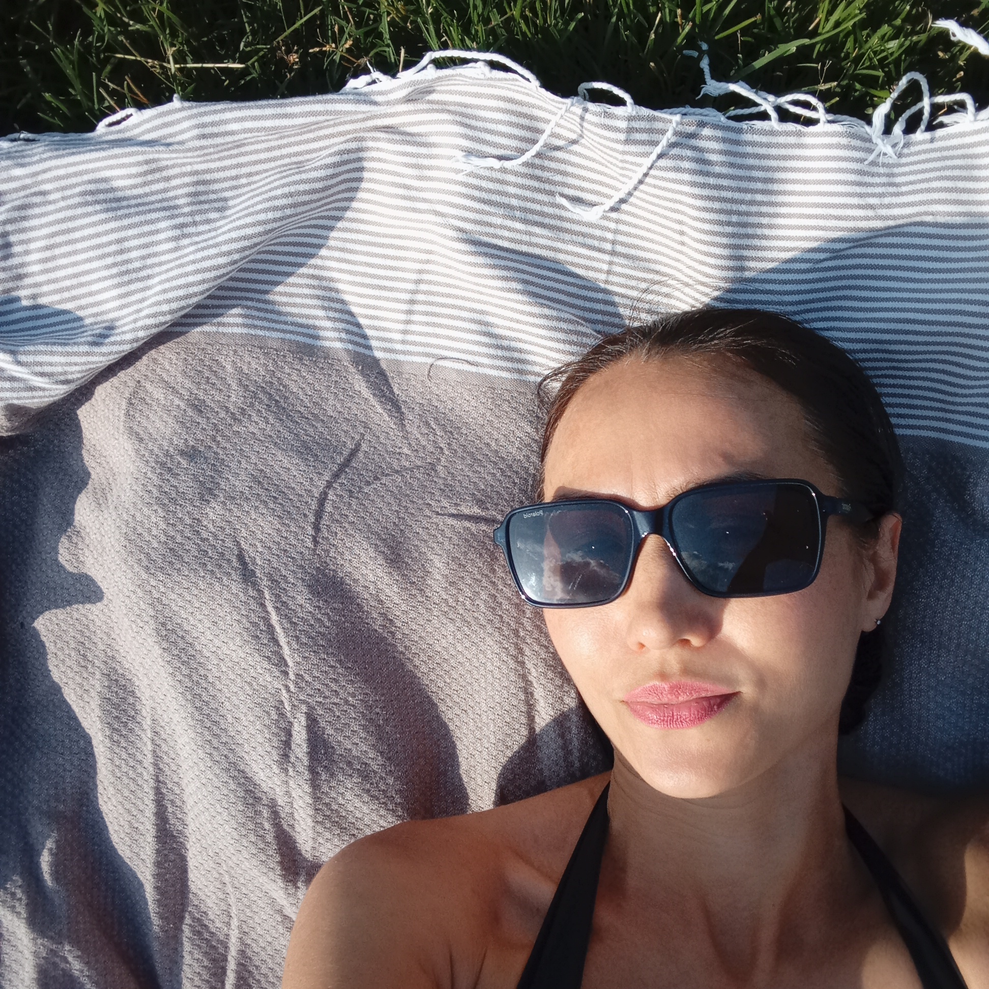 A close-up, overhead shot shows a woman with dark hair lying on a striped blanket and a textured grey blanket on a grassy area. She is wearing black swimwear and dark sunglasses. The scene is outdoors, with natural light casting shadows. The grass is green and appears to be a lawn. The striped blanket is white with grey stripes and has tassels. The grey blanket is textured and appears to be a heavier fabric. The woman's face is turned towards the camera, and she is wearing pink lipstick. The overall impression is of someone relaxing outdoors. The location is not specified in the image or the provided information.