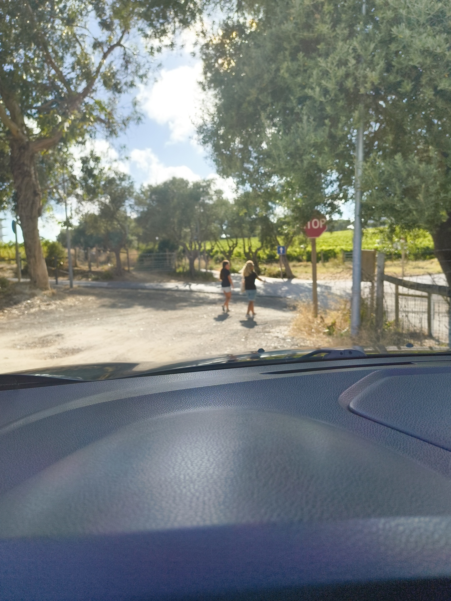 The image shows a view from inside a vehicle, looking out at an outdoor scene. The foreground consists of the textured surface of a car's dashboard and windshield. In the background, there is a dirt road with two young girls walking. One girl, on the left, wears a black top and shorts, and the other, on the right, wears a black top and denim shorts. A red octagonal stop sign with white lettering is visible on a pole to the right of the road. Trees and foliage line the sides of the road, with a glimpse of what appears to be a vineyard or agricultural fields further in the distance. The sky is blue with some clouds, and the sun is shining brightly. The scene appears to be in a rural or semi-rural setting, possibly on the outskirts of Oeiras, Portugal, given the typical landscape and vegetation found in the region.