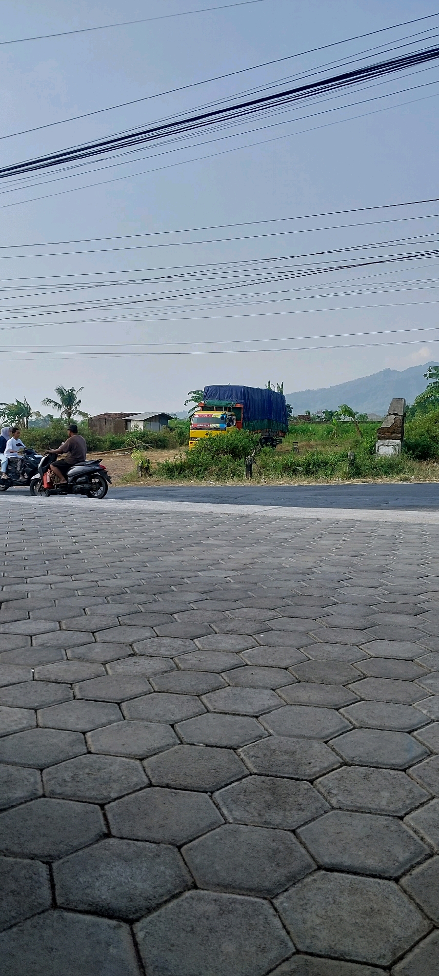 This image depicts a street scene in Keling, Indonesia, on a clear day. In the foreground, a hexagonal paver walkway leads towards a paved road. On the road, two motorcycles are traveling away from the viewer. The first motorcycle is ridden by a person wearing a dark shirt, with another person behind them. The second motorcycle, to the left, is occupied by two individuals, one in a white shirt and headscarf, and another person behind them. Beyond the road, a yellow truck with a blue tarp covering its cargo is parked on the side of the road. The background features a line of green vegetation, including trees and bushes, with several small buildings and a mountain range in the distance. Overhead, multiple power lines stretch across the sky.