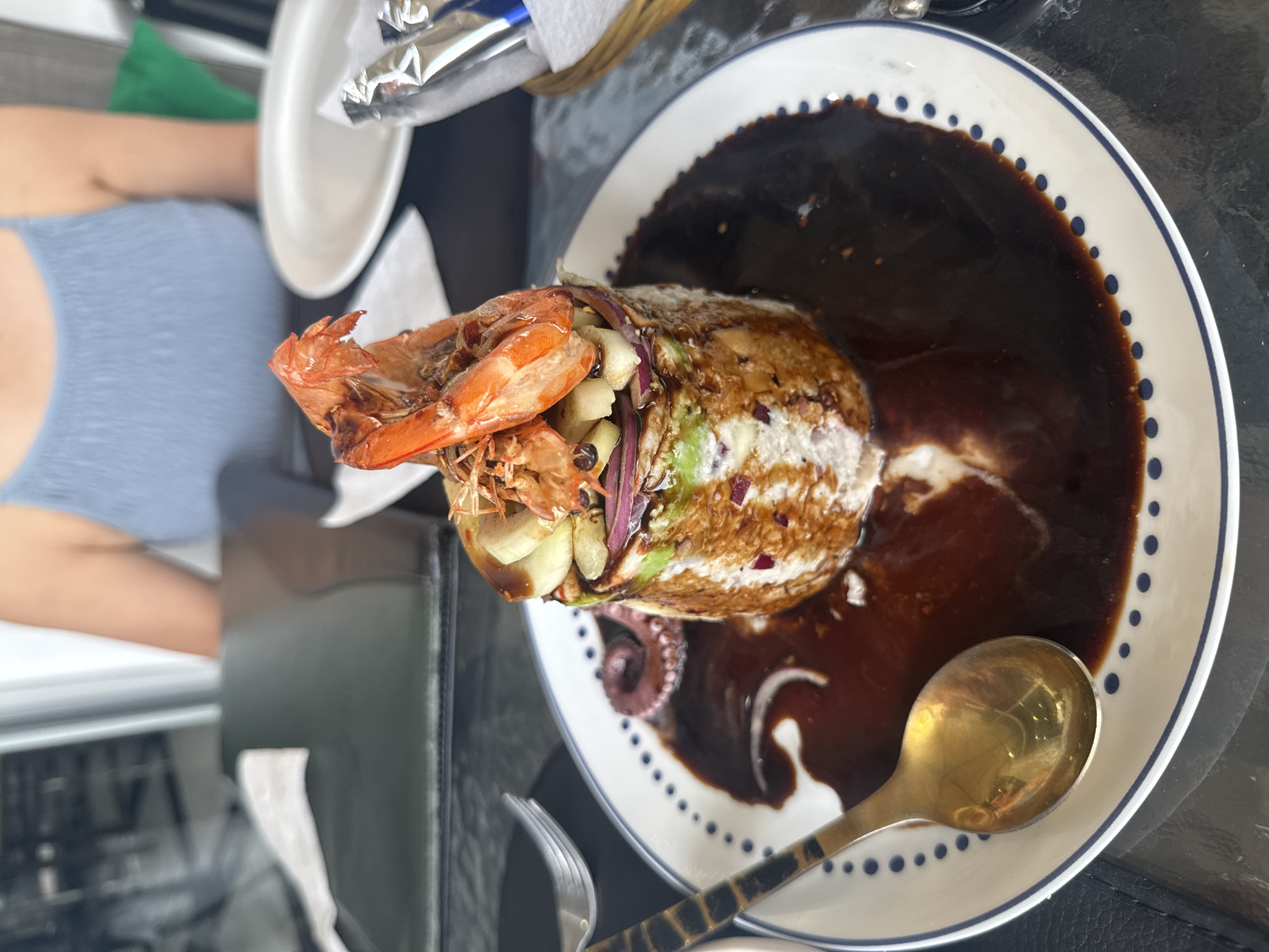 An overhead shot shows a seafood dish, likely served in Puebla, Mexico, on a white plate with a blue dot pattern rim. The dish consists of a mound of seafood, possibly a stuffed avocado or similar, topped with grilled shrimp. Around the base of the mound is a dark brown sauce, which might be a mole or similar traditional Mexican sauce. The plate has a spoon with a dark patterned handle, and a fork lies next to the plate. A partial view of a person wearing a light blue top is on the left side of the frame. There are other restaurant elements in the background like plates, foil-wrapped items, and a basket. The location is suggested by the style of the dish and the reference to Puebla, Mexico.