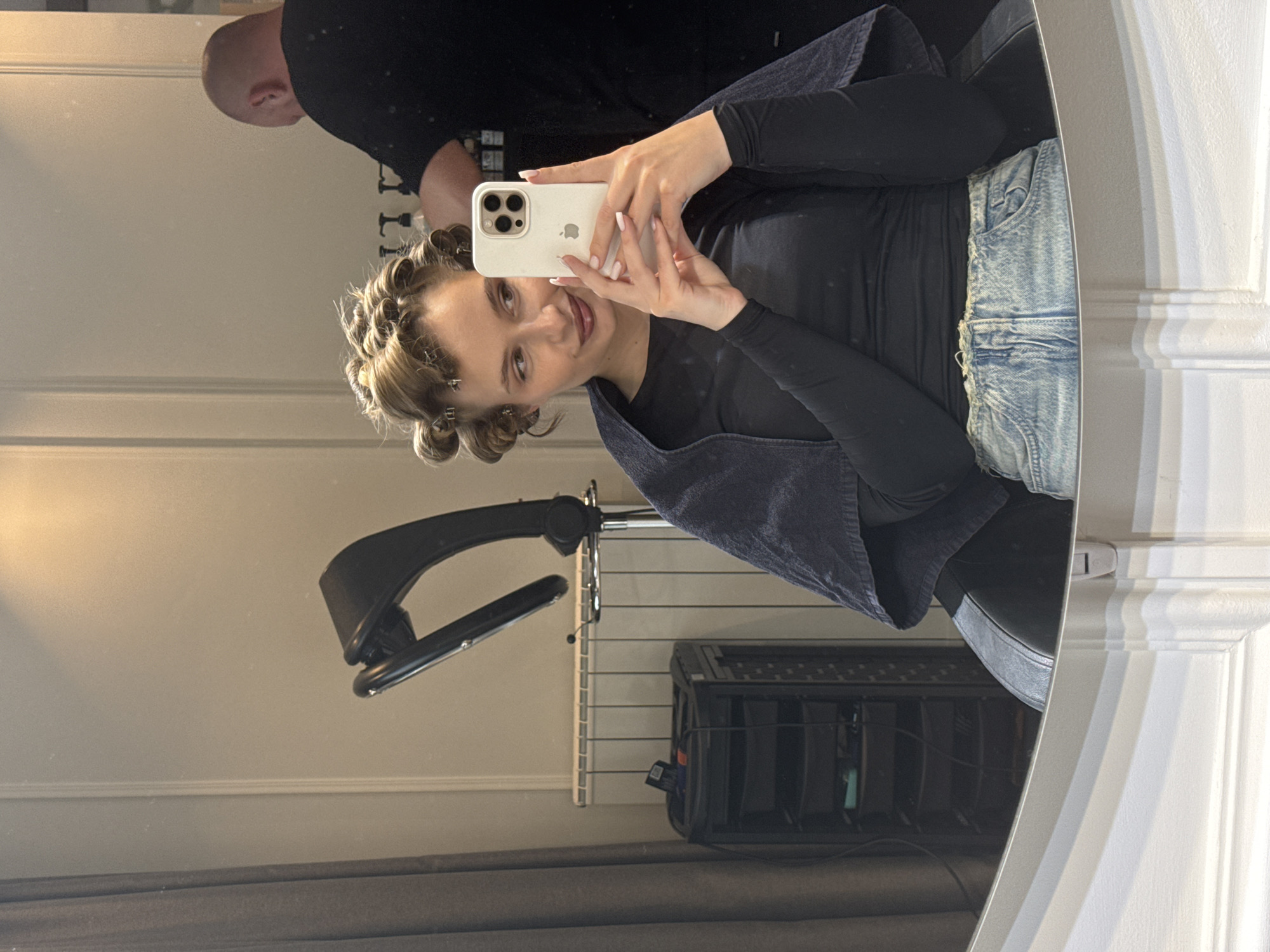 A woman with her hair in curlers is taking a selfie in a mirror. She is holding a white iPhone in her hands. She is wearing a black long-sleeved shirt and acid wash jeans. A blue towel is draped over her shoulders. A bald man is standing behind her, partially visible in the reflection.
In the background, there is a black hair dryer on a stand, a white wall, and a black roller case on the floor. The mirror has a white ornate frame.
The location is a room, likely a salon or private residence, in Belgrade, Serbia.