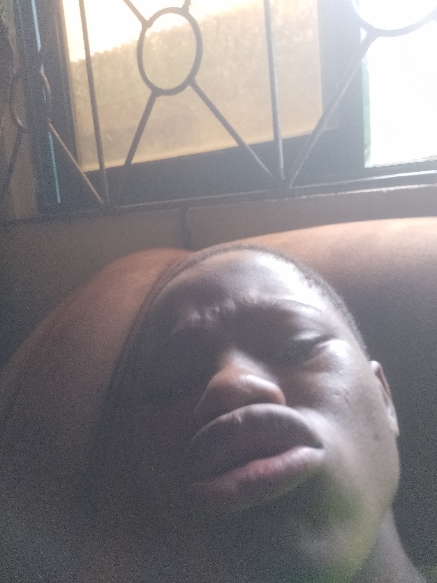 An image showing a person's face and upper body with a window in the background. The window has a metal frame with circular and linear designs. The person has dark skin, closed eyes, and slightly parted lips. The lighting is bright and diffused, making it difficult to discern fine details. The background suggests an interior setting, possibly within a home. No specific location in Shangisha, Nigeria can be definitively determined from the provided image alone.