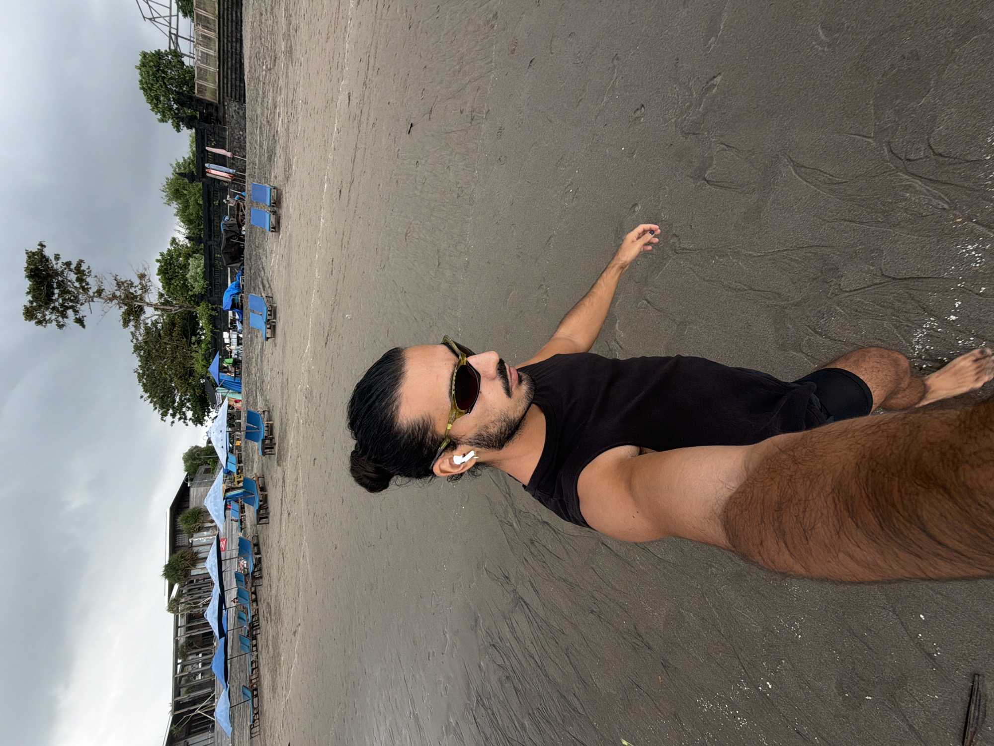 A man with dark hair pulled into a bun, wearing a black tank top, sunglasses, and white earbuds, is walking on a dark sand beach in North Kuta, Indonesia. He is holding a camera in his right hand, taking a selfie. In the background, there are rows of blue lounge chairs and umbrellas set up on the beach. Further back, there is a building with a modern architectural design, surrounded by trees. The sky is overcast with clouds.