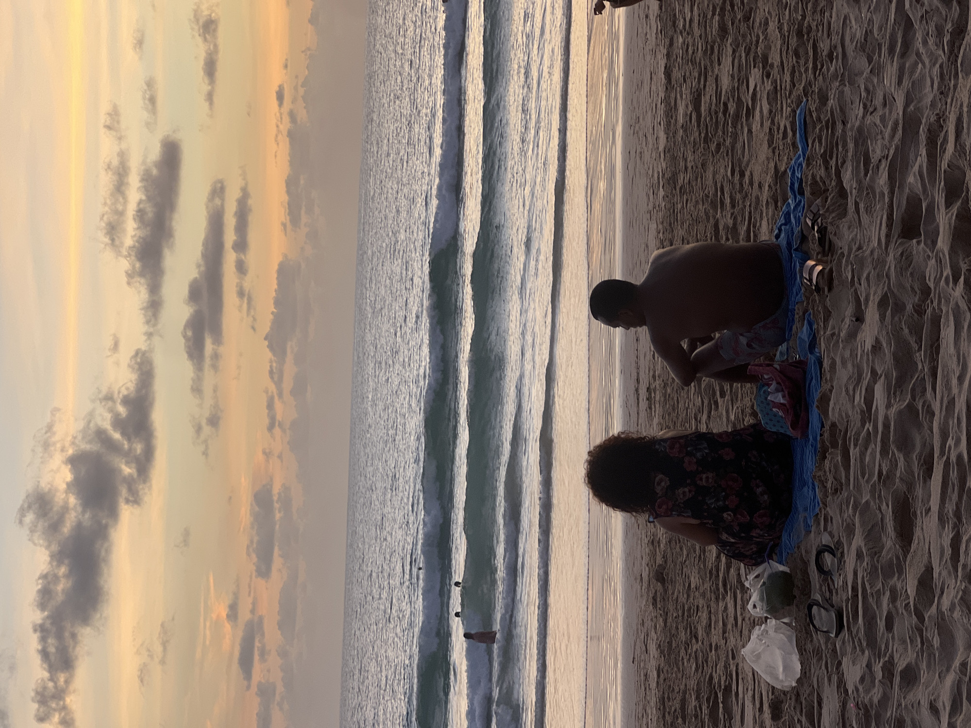 A couple sits on a beach in Choeng Thale, Thailand, facing the ocean as the sun sets. The man is shirtless and wearing patterned shorts, while the woman wears a floral print dress. They are seated on a blue towel on the sand. In the background, several people are swimming and surfing in the ocean. The sky is filled with soft clouds, reflecting the warm colors of the sunset. The sand is textured with footprints, and various items, including plastic bags, a coconut, and flip-flops, are scattered around the couple.