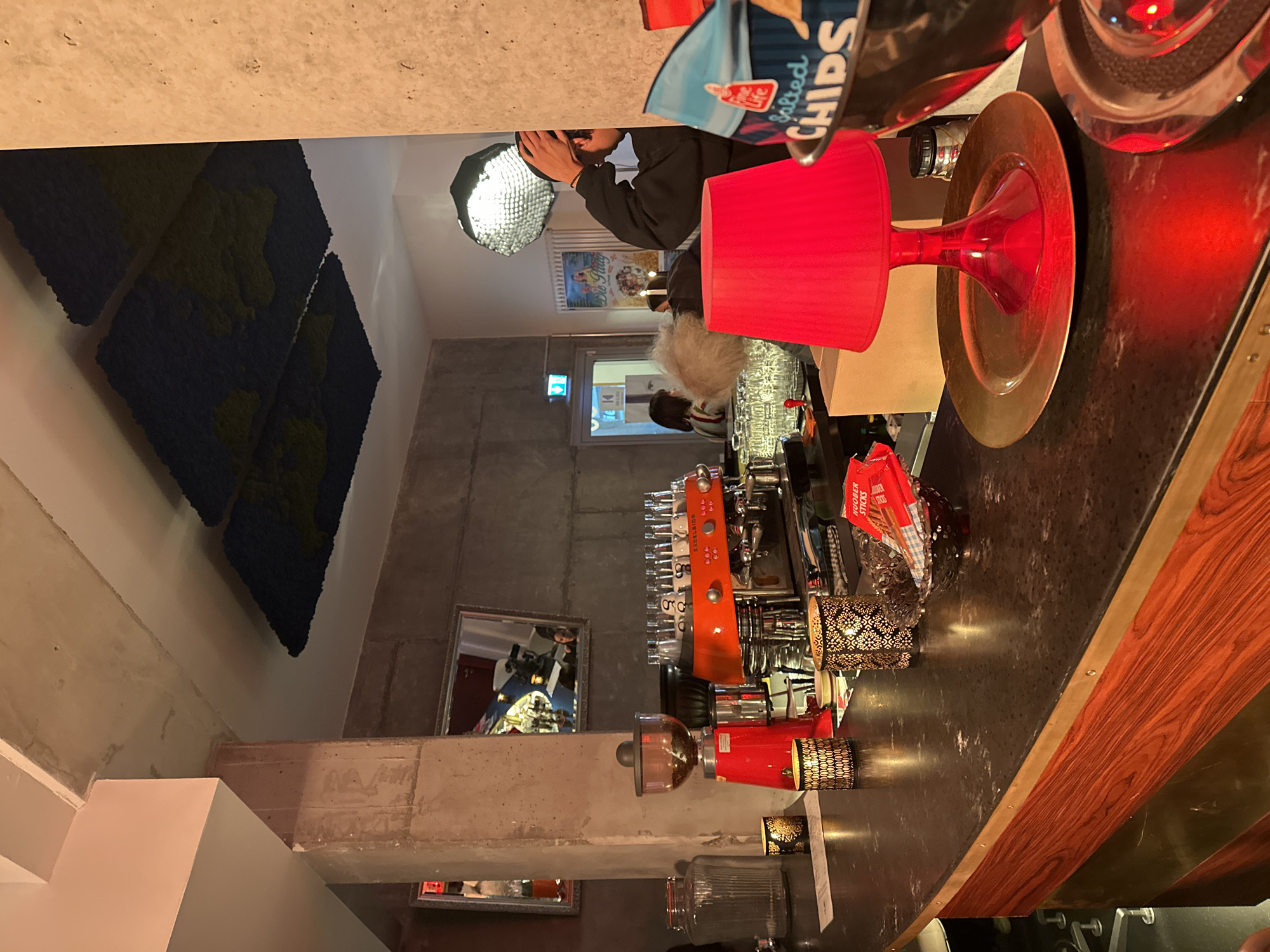 This image captures the interior of a modern bar or cafe, likely in the evening. The setting features an industrial-chic aesthetic with concrete walls and a long, dark counter.
Several people are visible. In the background, a person with dark hair and a mustache is holding a device, possibly taking a photograph or video, illuminated by a large, circular softbox light. Two other individuals are seated at the counter with their backs to the viewer; one has light-colored, fluffy hair, and the other has dark hair. Reflections in a mirror on the back wall show glimpses of other patrons and bottles behind the bar.
The counter is a focal point, adorned with several red lampshades and decorative metallic lanterns featuring intricate patterns, casting warm light. A prominent orange and silver espresso machine, possibly a "Gaggia Milano," is situated on the counter, surrounded by numerous small glasses. A gold-colored charger plate and a red glass vase also sit on the counter. A partially visible bag of "Salted CHIPS" and a red box labeled "HOOVER STICKS" are also present.
The walls are decorated with dark blue and green textured art pieces resembling landmasses. A vintage-style poster, with partially readable text including "Die Flucht," hangs on a distant wall. An "EXIT" sign with a running person symbol is visible above a doorway, common in European establishments. The overall atmosphere appears relaxed and social, typical of a casual evening gathering at a bar or cafe in an urban setting like Berlin.
