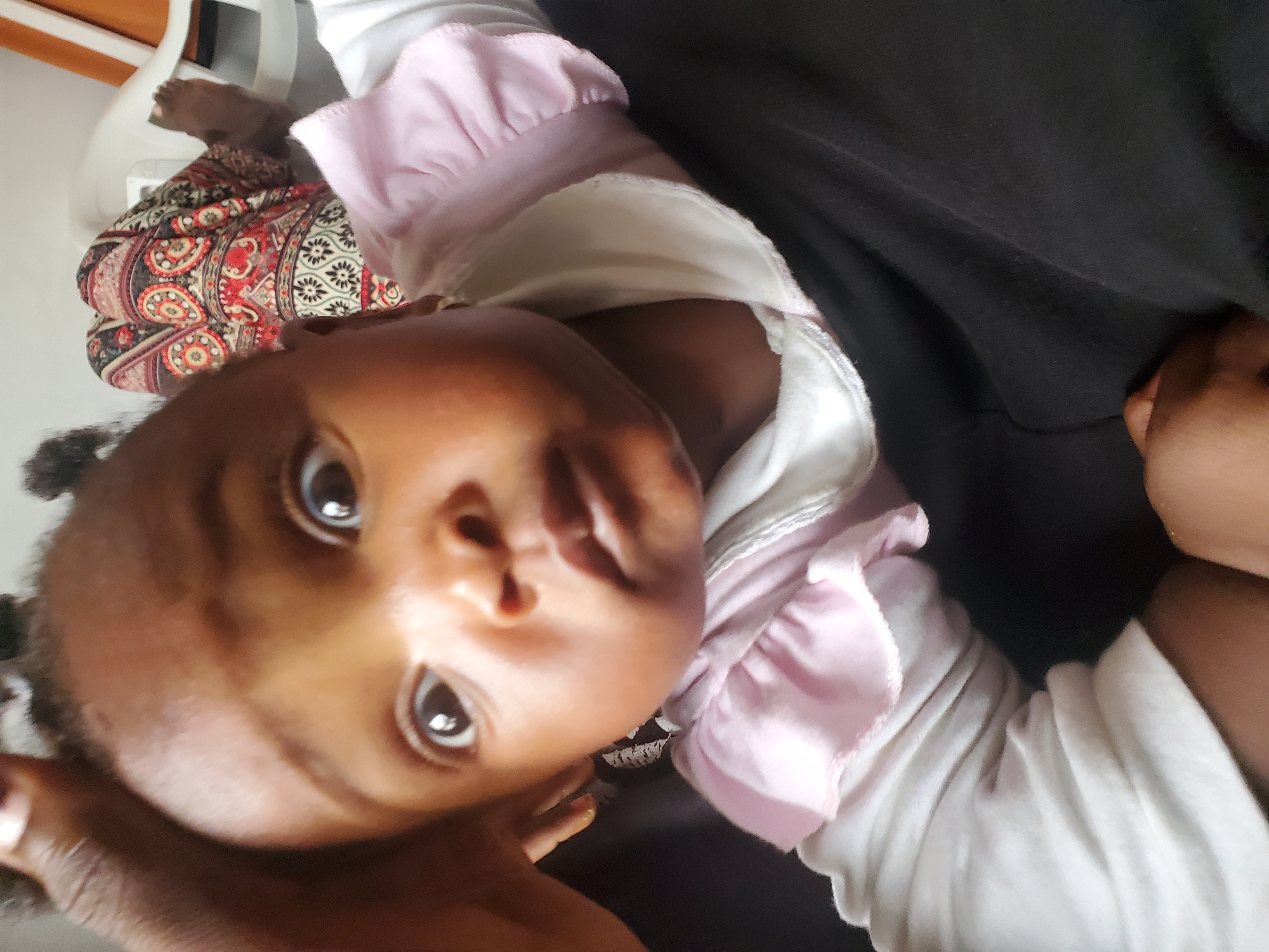 A close-up, low-angle, selfie-style shot features a young African infant in the foreground, looking directly at the camera with wide eyes and a slightly parted mouth. The infant is wearing a white long-sleeved shirt with a pink ruffled collar. The infant's head is tilted slightly to the right, and a dark hand is visible holding the infant's head from the left side of the frame.
In the background, slightly out of focus, a person is seated on a white plastic chair, wearing a colorful, patterned cloth. Part of another white plastic chair is visible to the right. The setting appears to be indoors, possibly a home. The overall lighting suggests an indoor environment with natural light. The image is taken from a perspective that emphasizes the infant's face. The location of Jos, Nigeria, cannot be determined from the visual information present in the image.