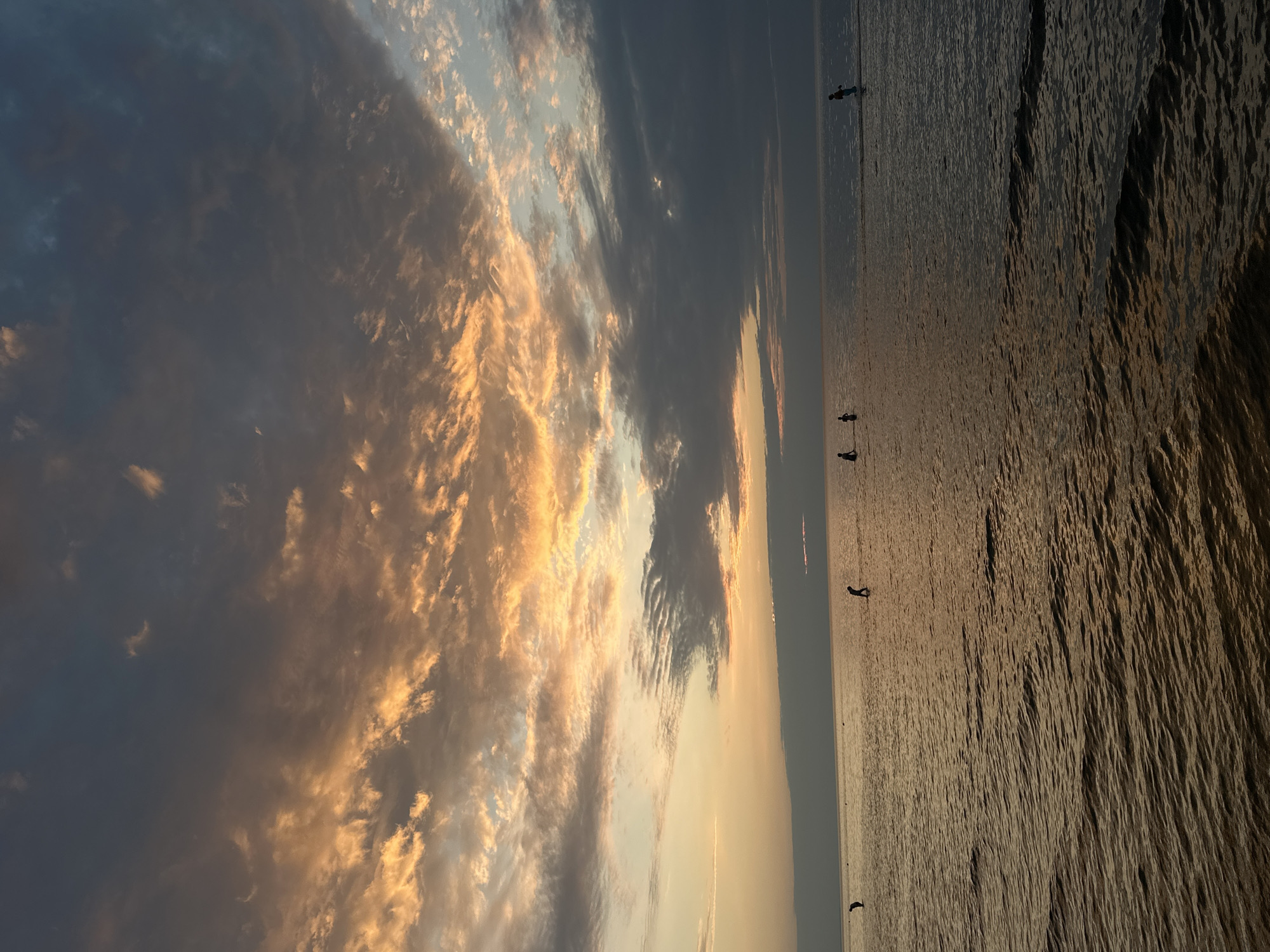 At sunset in Jūrmala, Latvia, the sky is a dramatic display of clouds tinged with orange and yellow, juxtaposed against darker, ominous gray formations. The vast expanse of the Baltic Sea reflects the sky's colors, with gentle ripples creating a textured surface. Silhouetted figures can be seen wading in the shallow water. One individual is standing further out, seemingly engaged in a water sport or activity. The overall impression is one of a tranquil evening at the seaside.