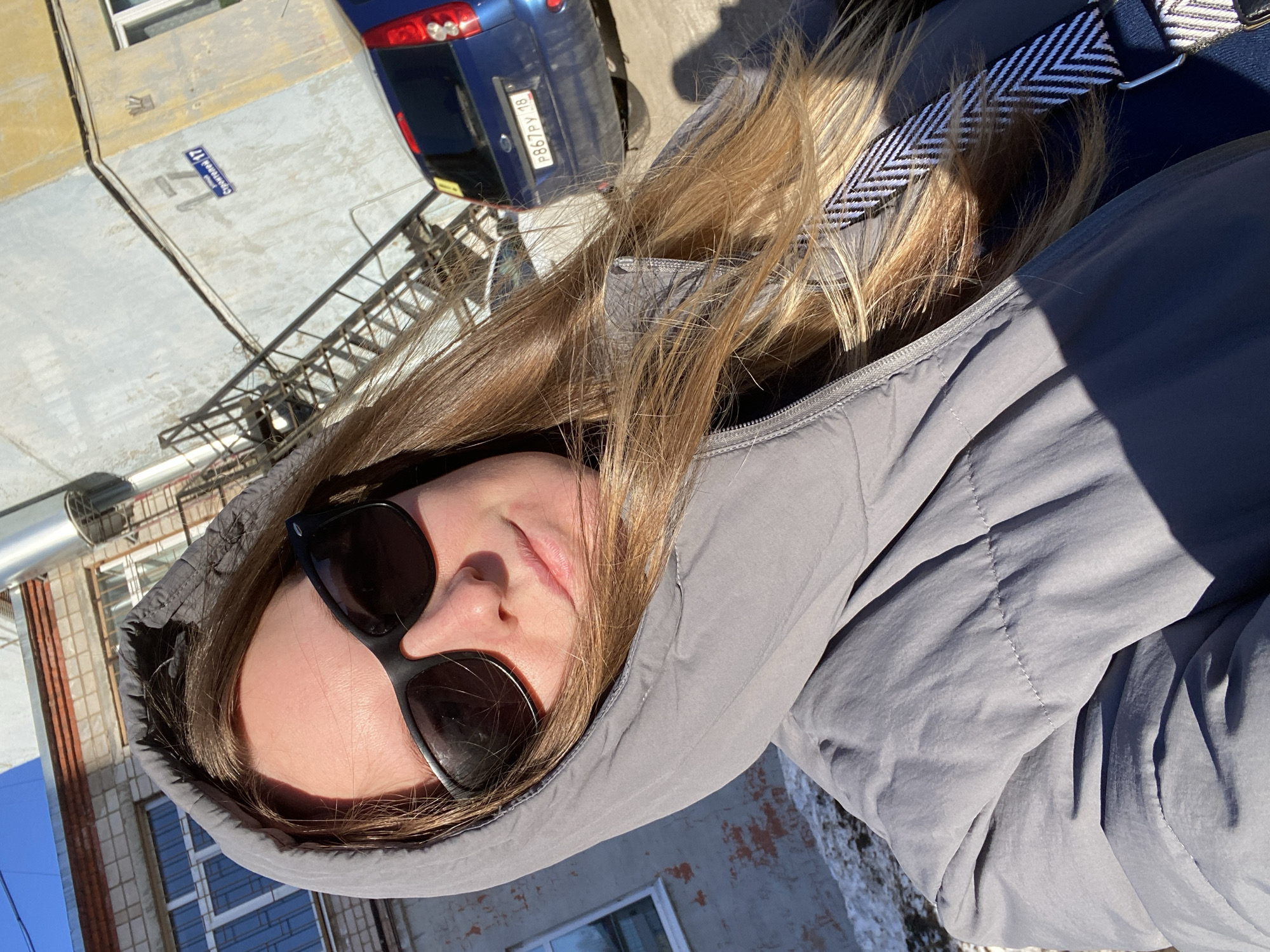 A selfie of a person with long, light brown hair, wearing sunglasses and a gray hooded jacket, taken outdoors on a sunny day. In the background are buildings, including one with red brick and white-framed windows. A blue car is partially visible in the upper left corner, with the license plate "F867PY 18". A street sign on a building indicates "Строителей 17". The location is likely the city of Noviy, Russia, based on the license plate information.