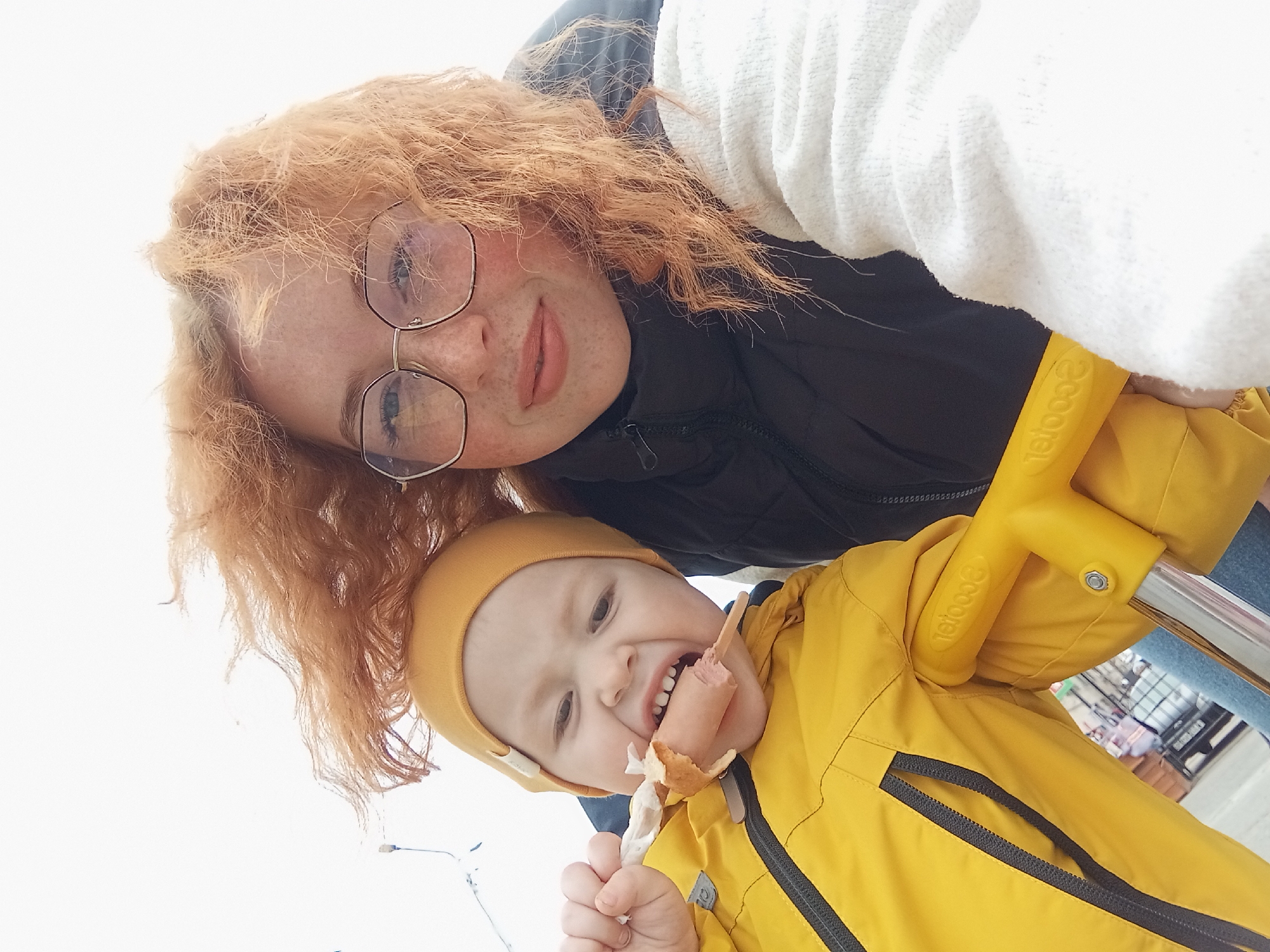 A woman with red curly hair and glasses is holding a child in a yellow coat and hat. The child is eating a sausage on a stick. They are outside, possibly in the city of Yurgamysh, Russia. A scooter handle is visible.
