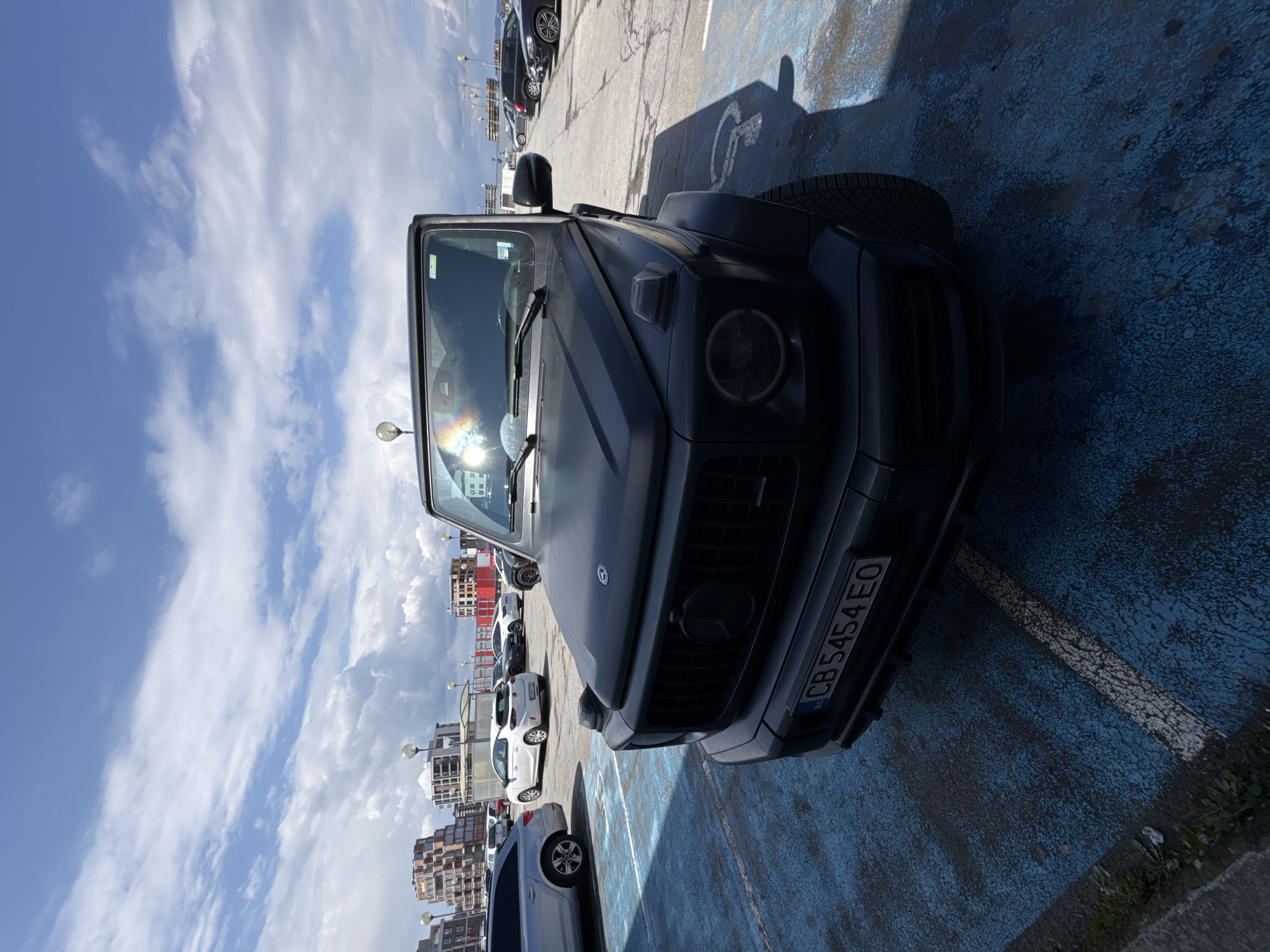 The image shows a black Mercedes-Benz G-Class parked in a blue parking space. The license plate reads "CB5454EO" and indicates the car is registered in Burgas, Bulgaria. Other cars are parked nearby, and in the background, buildings and apartment complexes are visible, suggestive of an urban or residential area. The sky is partly cloudy. The parking space appears to be painted blue.