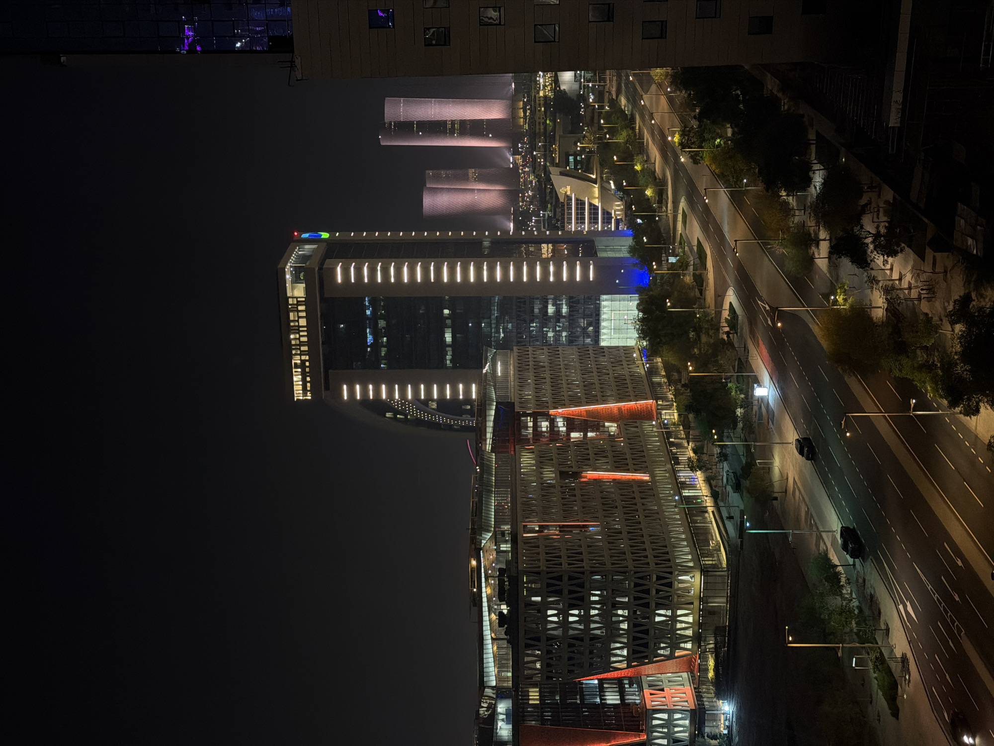 This is a nighttime aerial shot of a modern cityscape. In the foreground, a multi-lane road runs diagonally from bottom left to the middle right of the frame, with several cars visible. Streetlights illuminate the road and sidewalks. On the left side of the road, a large, angular building with a patterned facade and red and white lights is prominent. To the right of this building and further back, a tall, slender skyscraper with vertical white lighting stands out. In the background, several other illuminated skyscrapers rise, some with a distinctive cylindrical shape. Trees line the sides of the road and surrounding areas, and the overall scene is dark with artificial lights from buildings and street lamps.