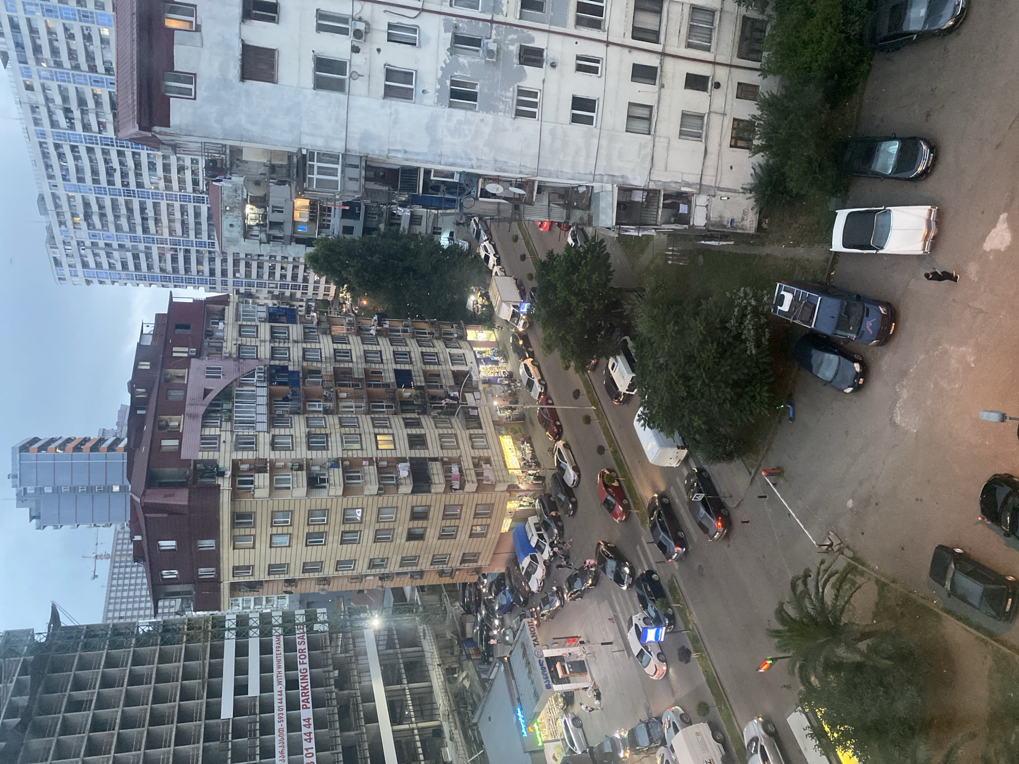 An overhead medium shot captures a bustling city street scene with multiple apartment buildings and various vehicles. A large, unfinished building with exposed concrete framing stands on the left, adorned with a banner advertising "PARKING FOR SALE." Adjacent to it, a multi-story apartment building with a reddish-brown mansard roof features numerous windows and balconies.
In the foreground, a street is lined with parked cars and a few moving vehicles. A gas station with a blue and yellow sign reading "TACHTOIL" is visible on the left, with a few cars refueling. Further down the street, more cars are parked along the roadside.
Several other apartment buildings of varying heights and architectural styles populate the background, some with modern glass facades and others with older, more weathered exteriors. The sky is overcast, suggesting it might be late afternoon or early evening. The scene appears to be a dense urban environment with a mix of residential and commercial activity.