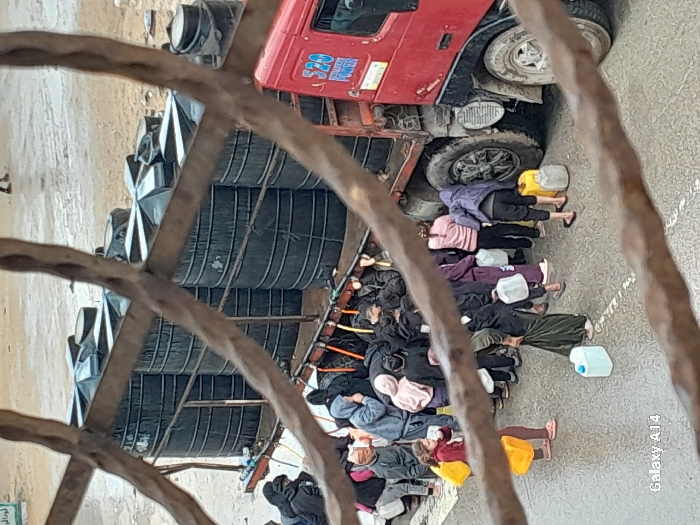 The image depicts an elevated view of a crowd gathered around a water distribution truck in Khan Yunis, Palestinian Territory. A red truck with a flatbed carries at least ten black, cylindrical water tanks. Approximately 20-30 individuals, including adults and children, are clustered around the truck's side, actively engaged in filling various containers with water. Many people hold clear, white, or yellow plastic jerrycans and bottles. One young girl in a red top and yellow skirt holds a yellow bag. The ground is wet asphalt or packed dirt, with visible puddles, suggesting recent rain or water spillage. In the background, an open, dusty area is visible, with a faint sign displaying Arabic script in the upper left. The scene is partially obscured by blurred, dark, organic-looking elements in the foreground, likely a window grille or fence.