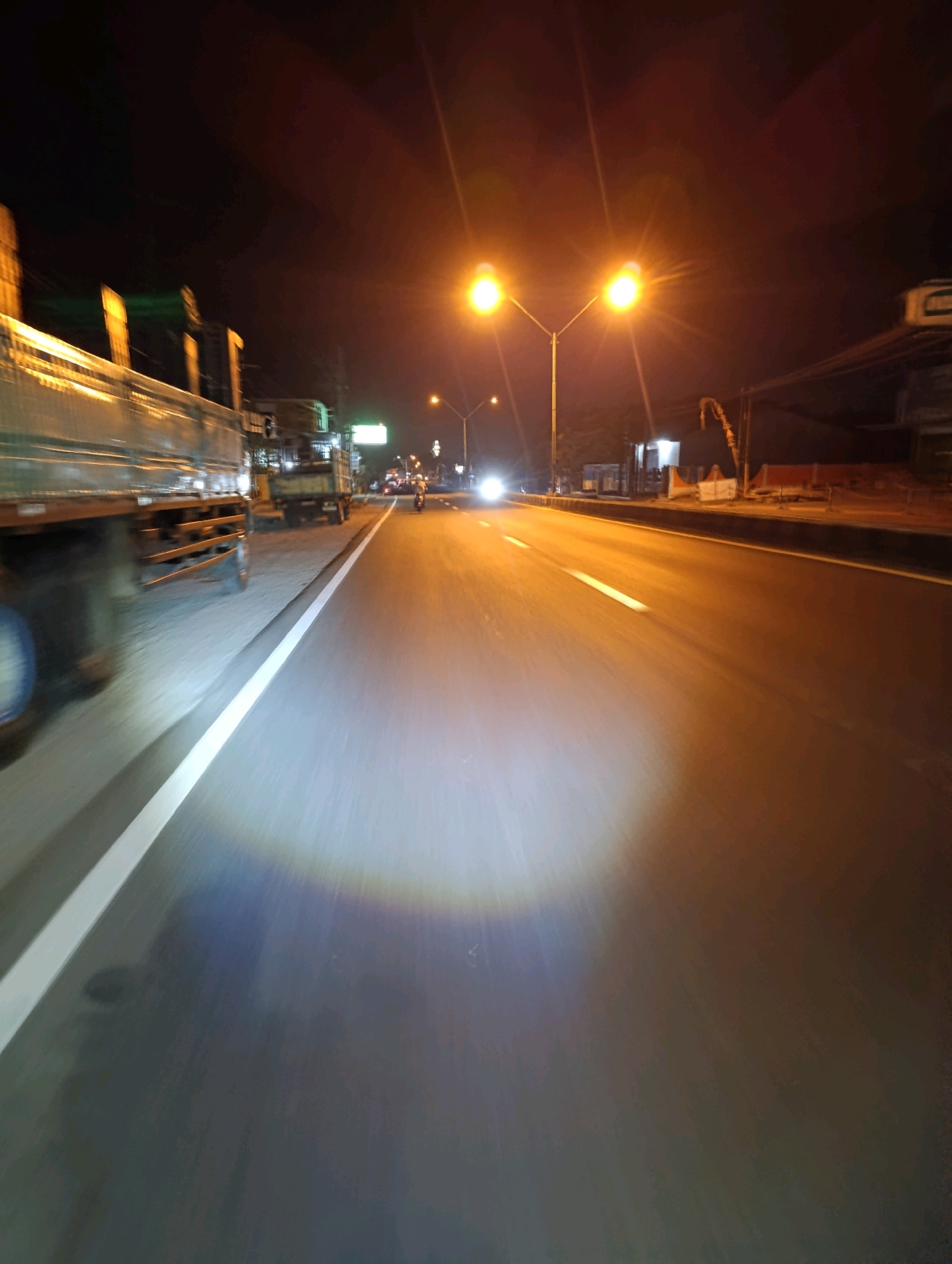 The image captures a nighttime scene on a paved road in Glagah Sari, Indonesia. The perspective appears to be from a moving vehicle, likely a motorcycle or bicycle, given the low-angle viewpoint and the illuminated circle on the road ahead, suggesting a headlight. On the left side of the road, the side of a large truck is visible, its metallic surface reflecting the ambient light. The truck's wheels are blurred, indicating motion. Beyond the truck, other vehicles and structures are dimly lit. In the distance, two bright streetlights illuminate a section of the road, casting a yellow glow. Further down the road, a single motorcycle is visible, its headlight also illuminated. The road itself has white lane markings, and the asphalt surface is textured. The background is dark, with hints of trees and buildings silhouetted against the night sky. The overall impression is that of a typical urban or suburban street at night, with the usual elements of traffic and street lighting.
