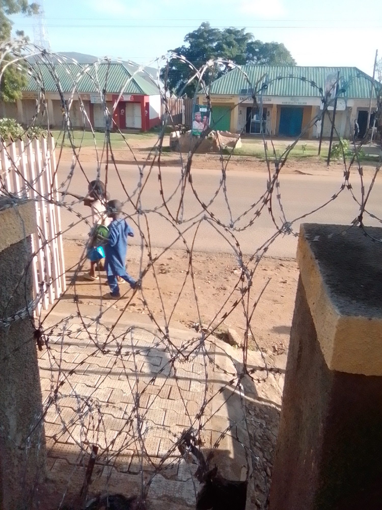 The image shows a street scene in Galadima, Nigeria, with two children walking on a paved area near a road. The foreground is dominated by a barbed wire fence, creating a visual barrier. Behind the fence, a red and white building with a green, ribbed roof is visible, appearing to be a shop or business. Another building with a similar green roof is to the left, with signs suggesting it's a commercial establishment. The children, one wearing a blue outfit and carrying a green object, are walking away from the camera. The environment appears dry and dusty, with some vegetation visible in the background.
