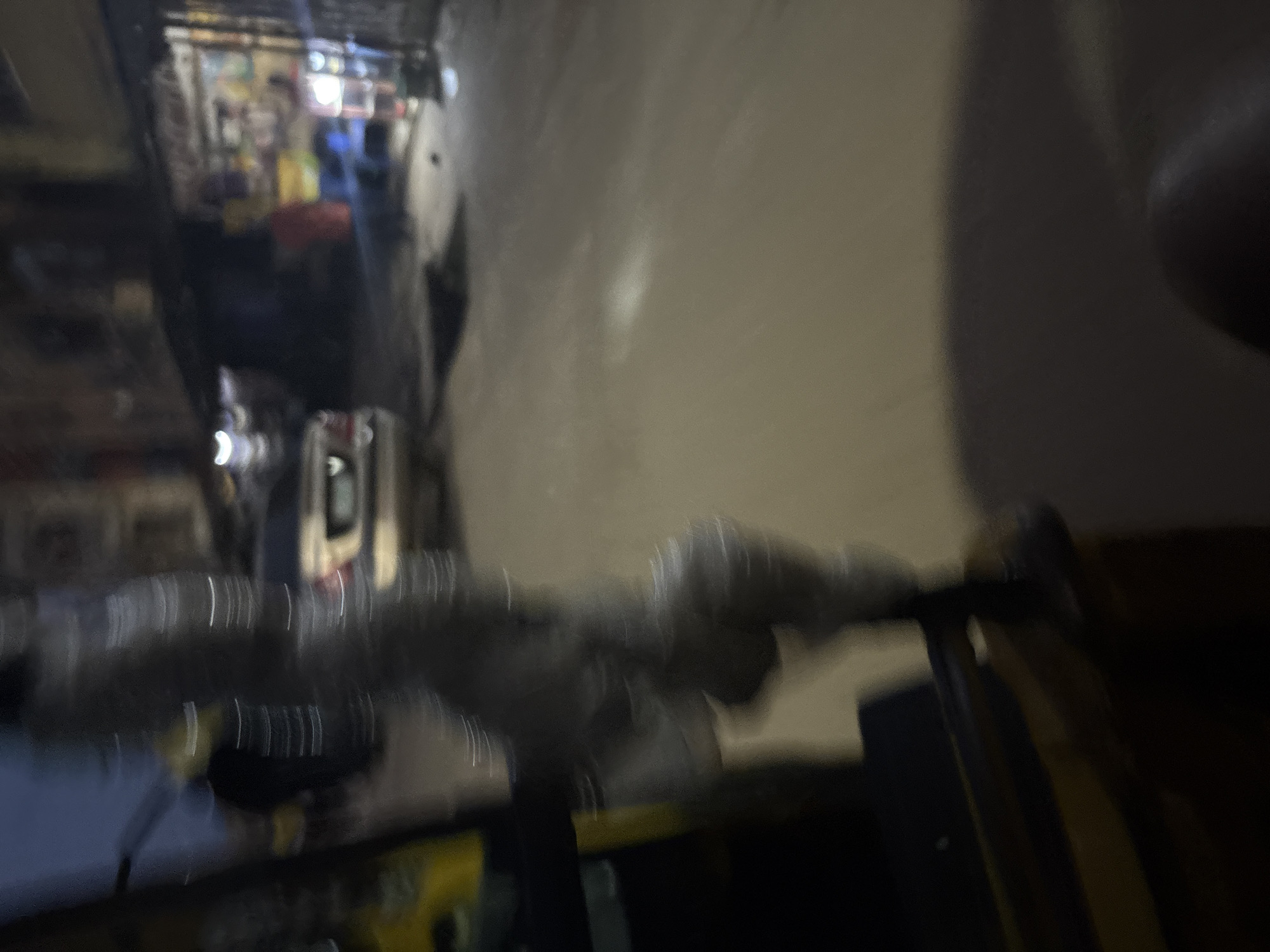 The image depicts a blurry street scene at night, likely in Onitsha, Nigeria. In the foreground, a dark, textured object, possibly a metal pole or railing, dominates the left side of the frame. To the left of this object, a yellow and black structure is partially visible, suggesting it might be part of a vehicle or a temporary stall. In the background, a white car is parked on the right side of the street. Beyond the car, buildings line the street, with visible shop fronts displaying various items, including what appear to be bottles and other products. The street itself is paved and dimly lit, with some scattered debris visible on the ground. The overall impression is one of a bustling, albeit somewhat disorganized, urban environment at night.