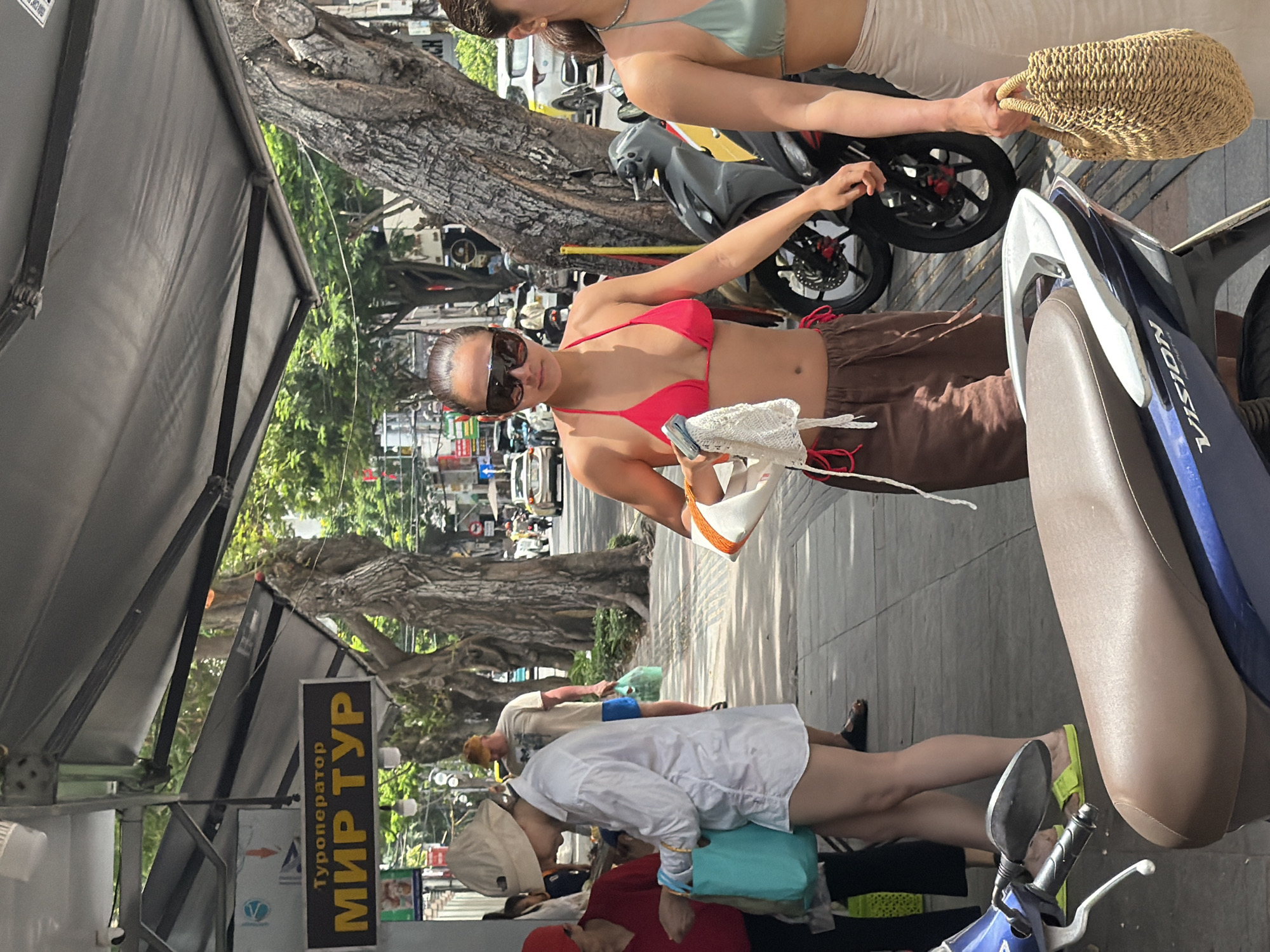 The image shows a sunlit street scene in Nha Trang Ward, Vietnam. A woman in a pink bikini top and brown shorts holds a white crocheted hat. To her right is another woman in a light green top and beige shorts, holding a woven bag. Behind them, two motorcycles are parked on the sidewalk. Beneath a black awning, people are standing near a travel agency sign in Russian: "Typonepatop Mup Typ." Other pedestrians, wearing casual clothing, pass by in the background. Trees line the street, creating dappled sunlight.