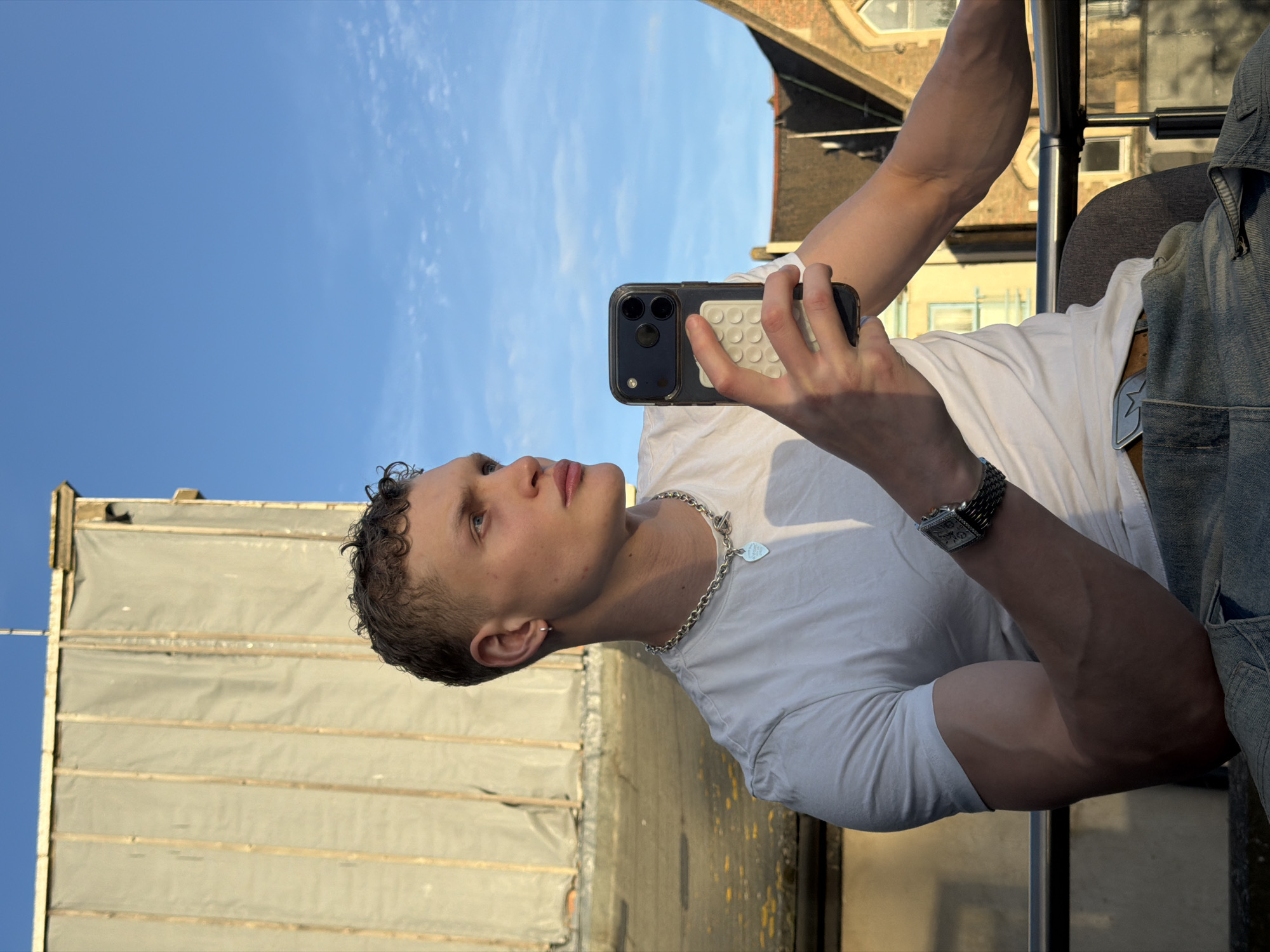 A young, light-skinned man with short, curly dark hair is holding a smartphone in his right hand, apparently taking a photo. He's wearing a white t-shirt, a silver chain necklace with a heart pendant, and a wristwatch. He appears to be on a double-decker bus. Behind him and to the left is a building facade covered with tarpaulin. In the background on the right is another building with a pitched roof and windows. The sky is blue with scattered clouds. Based on the architecture and bus infrastructure, the location is likely in London, United Kingdom.