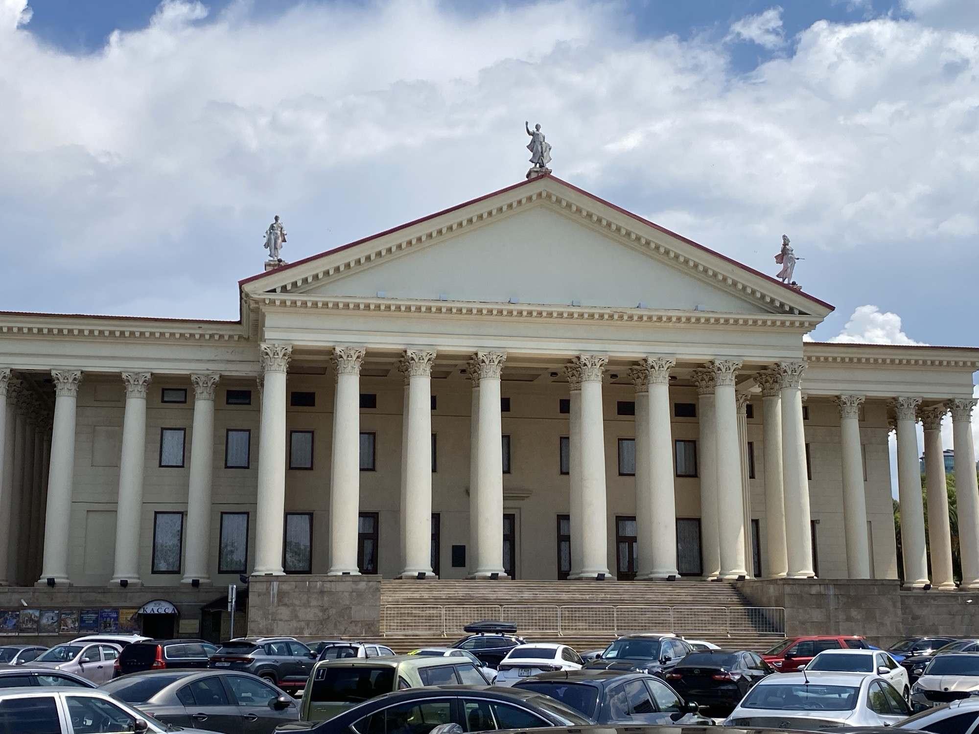 This image depicts a large, neoclassical building with a grand facade featuring numerous tall, white columns and a triangular pediment at the top. Statues of figures adorn the roofline and the pediment. Below the colonnade, there are several windows and a visible entrance with a sign that reads "КАССА" (KASSA), indicating a ticket office or box office.
In front of the building, a parking lot is filled with a variety of cars, suggesting that people are either arriving at or departing from the building for an event or visit. The scene appears to be set during the daytime, with bright sunlight casting shadows, indicating it's likely midday or afternoon. The sky is partly cloudy, with large white clouds scattered across a blue sky.
The architectural style, with its columns and pediment, hints at a public building, possibly a theater, opera house, or government institution. The presence of the ticket office further reinforces the idea of a place of entertainment or public access. The overall impression is one of a significant and established civic structure. The location context provided, Sochi, Russia, adds to the understanding of the setting.
