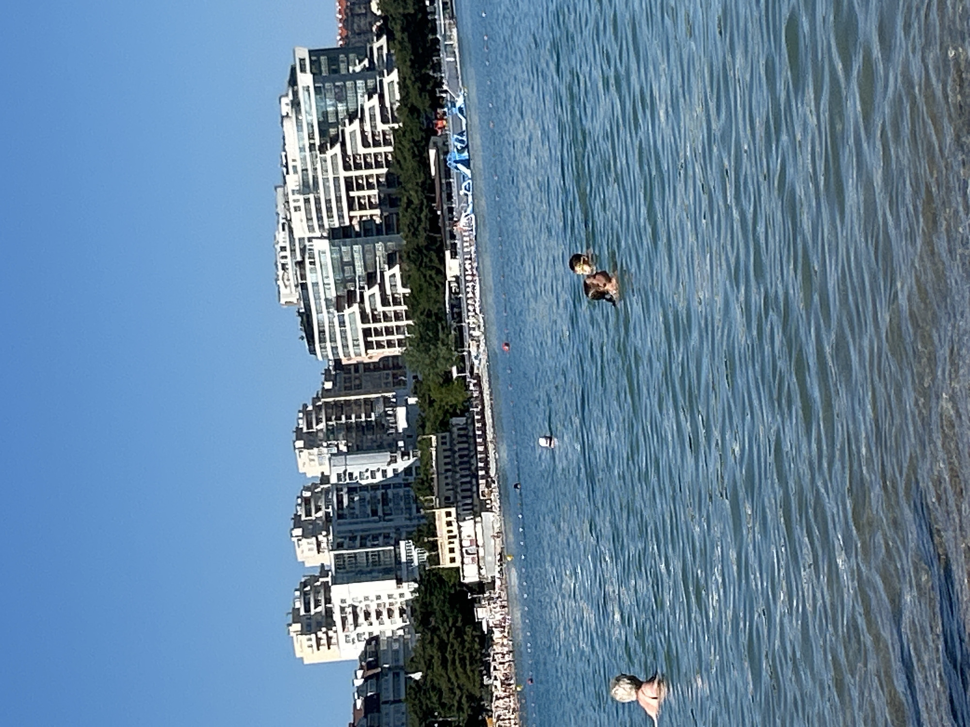 The image depicts a sunny day at a beach in Gelendzhik, Russia, with the Black Sea in the foreground and a cityscape in the background. The water is a clear blue with gentle waves. In the foreground, a woman with blonde hair is swimming, facing away from the camera. Further out in the water, two children are swimming together; one is wearing goggles. A white buoy floats in the water to the right of the children.
Along the coastline, there is a beach with numerous umbrellas and beachgoers. Behind the beach, a white building with a distinctive architectural style stands out. Beyond that, a dense line of green trees separates the beach area from a series of modern, multi-story apartment buildings that dominate the cityscape. Some of these buildings have a stepped or terraced design, with balconies and many windows. The sky is a clear, bright blue, indicating fair weather. The overall scene is one of a bustling summer day at a seaside resort.