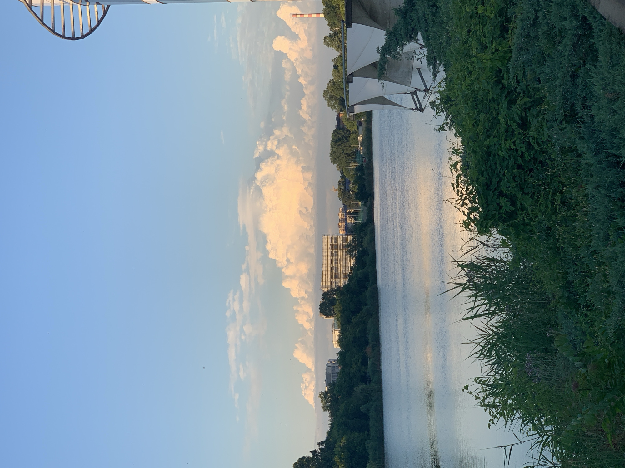 This is a photograph of a tranquil waterscape at sunset. The scene is dominated by a wide body of water, reflecting the soft hues of the sky. On the right side of the frame, lush green foliage and reeds line the bank, creating a natural border. In the distance, a cityscape emerges, with buildings partially obscured by trees. A prominent feature on the horizon is a tall, striped smokestack, suggesting an industrial presence. The sky is a blend of soft blues and warm oranges and yellows, indicative of a sunset. Cumulus clouds drift across the sky, catching the golden light. The overall mood is peaceful and serene, with the gentle light and calm water contributing to a sense of tranquility. The image is oriented vertically.