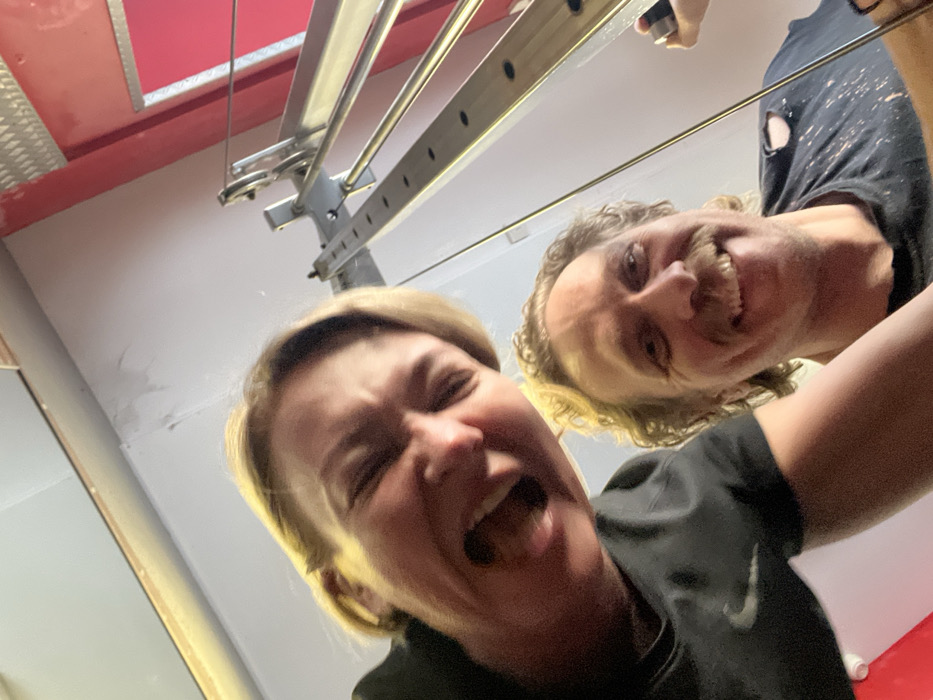 A low-angle selfie captures two individuals indoors, likely in a fitness setting. The person in the upper left, a female with blonde hair, is mid-laugh, mouth wide open, and eyes squinted, wearing a black top with a white Nike swoosh. The person below and to the right, a male with curly light brown hair and a mustache, smiles broadly, wearing a dark t-shirt with light splatters and a tear on the right shoulder. In the background, a multi-component metal structure, consistent with gym equipment like a cable machine, extends vertically, featuring adjustment holes and reflective surfaces. A bright red panel with a metallic, possibly ribbed, section is visible on the far right. The background walls are a plain light color. The precise location within Saint Petersburg, Russia, cannot be determined from the visual information available.