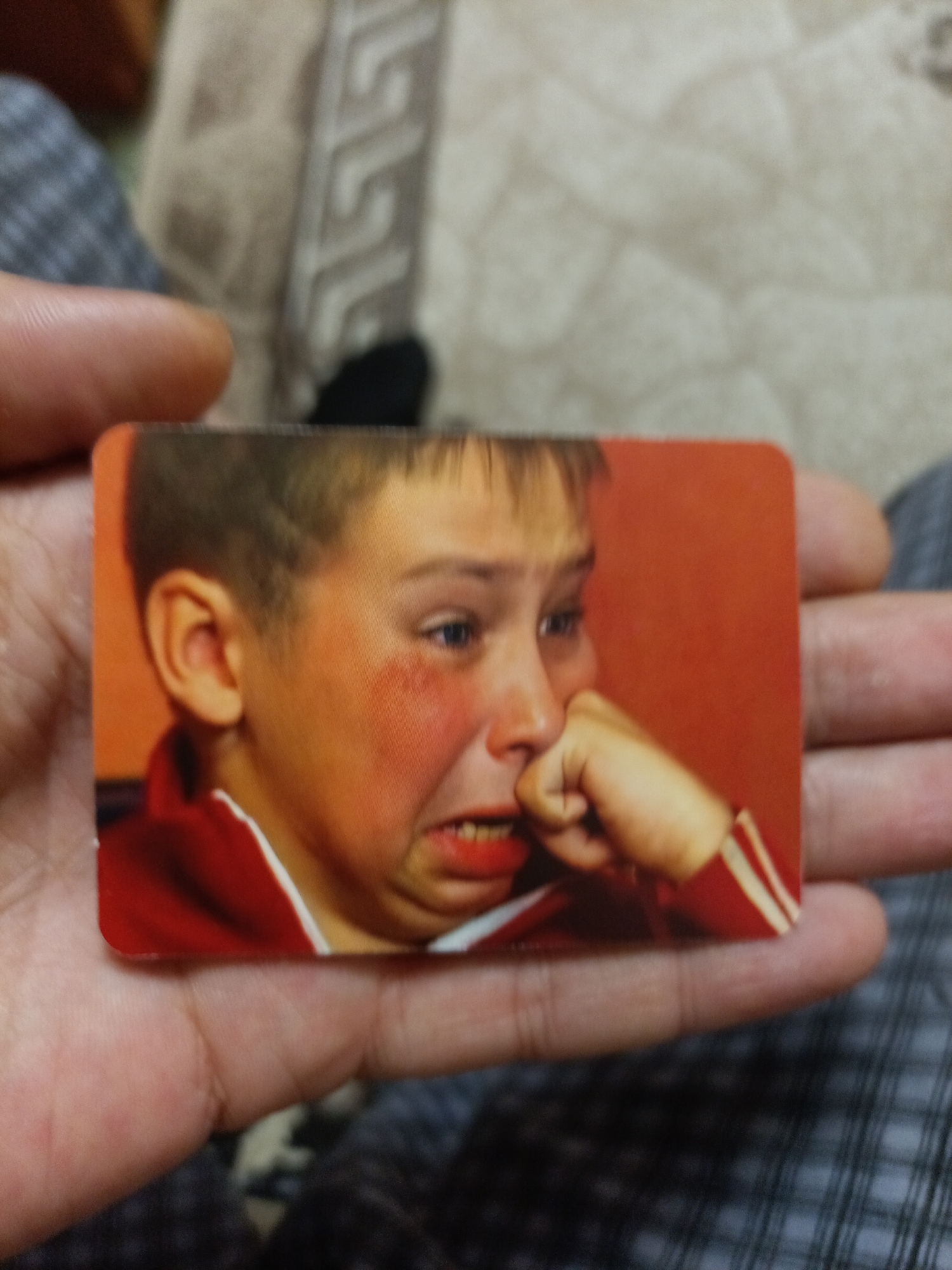 A close-up shot shows a hand holding a postcard featuring a young boy crying. The boy has red cheeks, his mouth is open in a sob, and his fist is pressed against his face. He is wearing a red jacket with white trim. The background of the postcard is a solid orange color. The hand holding the postcard appears to be an adult's, with visible fingerprints and skin texture. The surrounding environment is out of focus, but appears to be an indoor setting with patterned fabric in the background. There is no information or visual evidence in the image that suggests a specific location in the city of Nyagan, Russia.