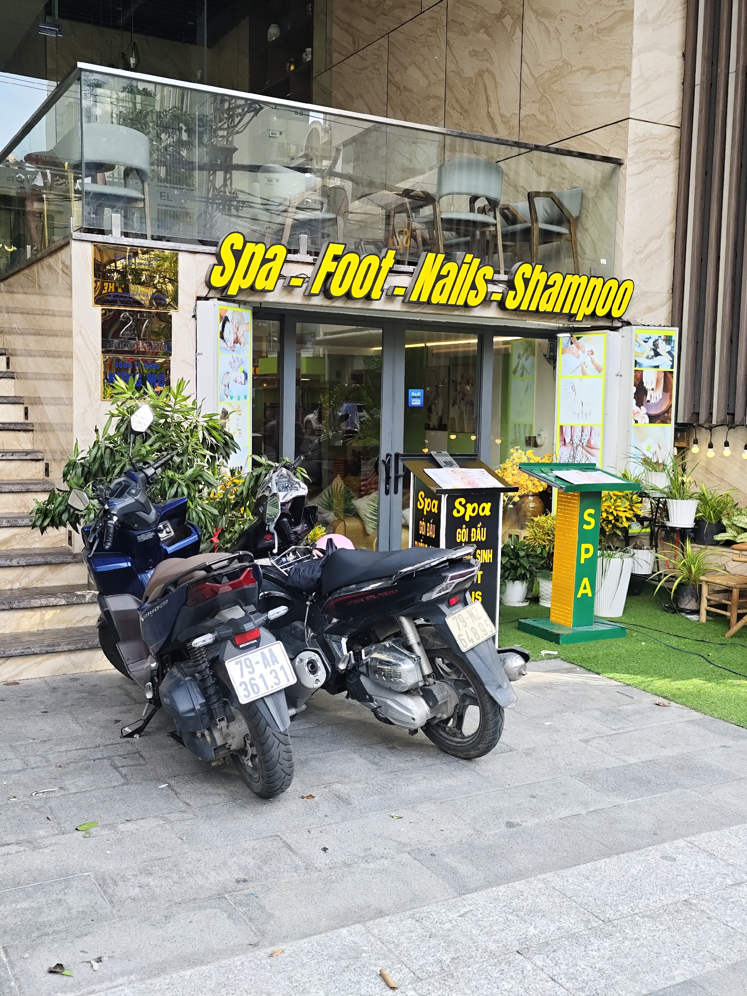 In the city of Nha Trang Ward, Vietnam, a street-level view captures the facade of a business identified by a bright yellow sign that reads "Spa - Foot - Nails - Shampoo". The business is situated on the ground floor of a building with a polished marble exterior and a glass-enclosed upper level displaying chairs. Two motorcycles are parked prominently on the sidewalk in front of the spa. The motorcycle closest to the viewer is dark blue and black, with a license plate reading "79-AA 361.31". The second motorcycle, mostly obscured by the first, is black and has a license plate that is partially visible. A lush green plant is positioned between the two motorcycles, and the ground is paved with grey stone tiles. To the right of the spa entrance, a green and yellow display stand with the letters "S.P.A." vertically arranged is placed on an artificial grass surface, surrounded by potted plants and a small wooden stool. The scene appears to be a typical street view of a commercial establishment in Vietnam.