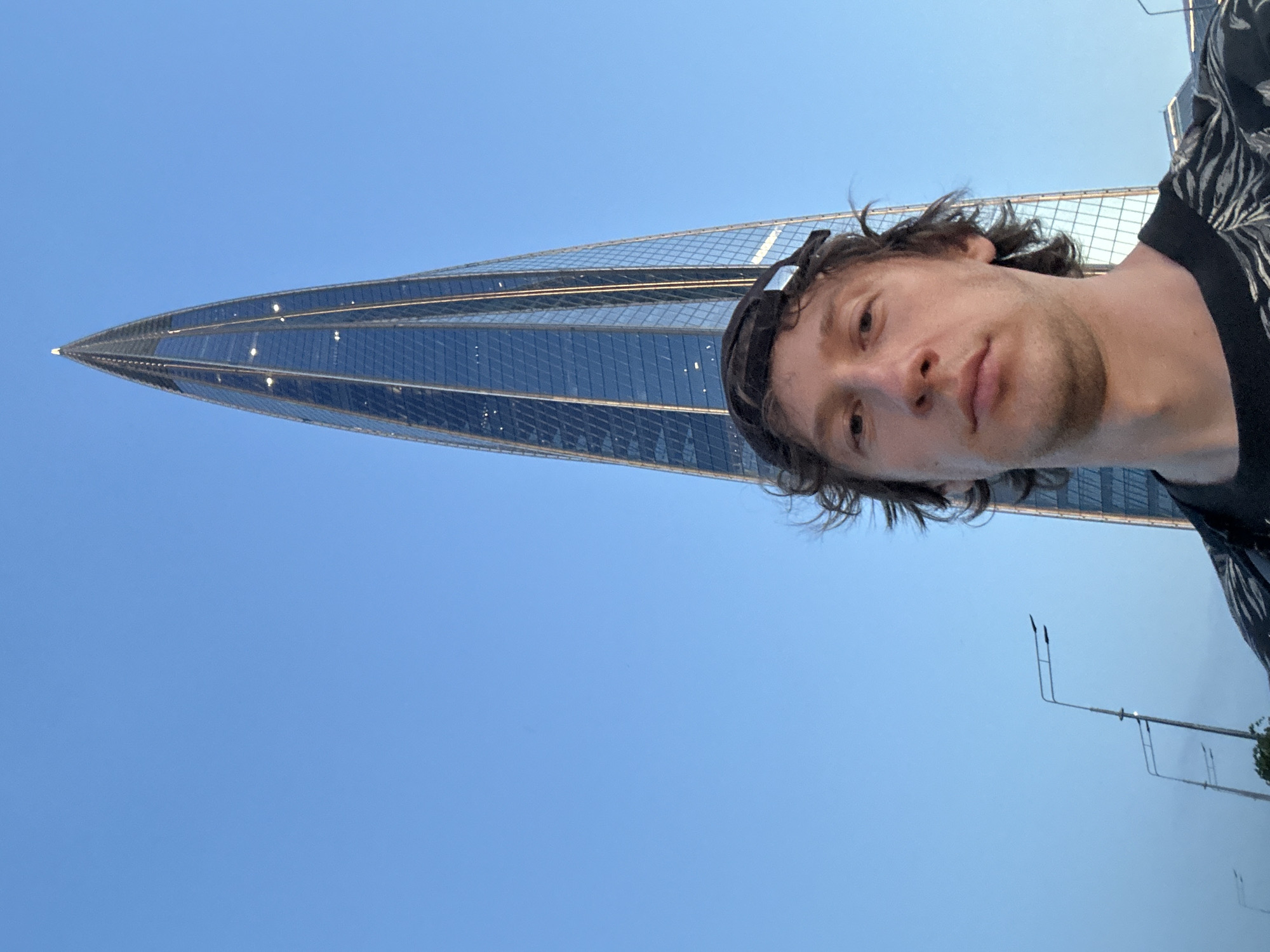In this image, a man wearing a baseball cap and a black shirt with a white pattern is taking a selfie in front of a very tall, sleek skyscraper. The skyscraper has a distinctive pointed top and is covered in reflective blue glass panels. The sky behind the building and the man is a clear, bright blue. In the foreground, below the man, are streetlights and what appears to be greenery, suggesting an outdoor urban setting. The skyscraper is the Lakhta Center, a landmark in Saint Petersburg, Russia.