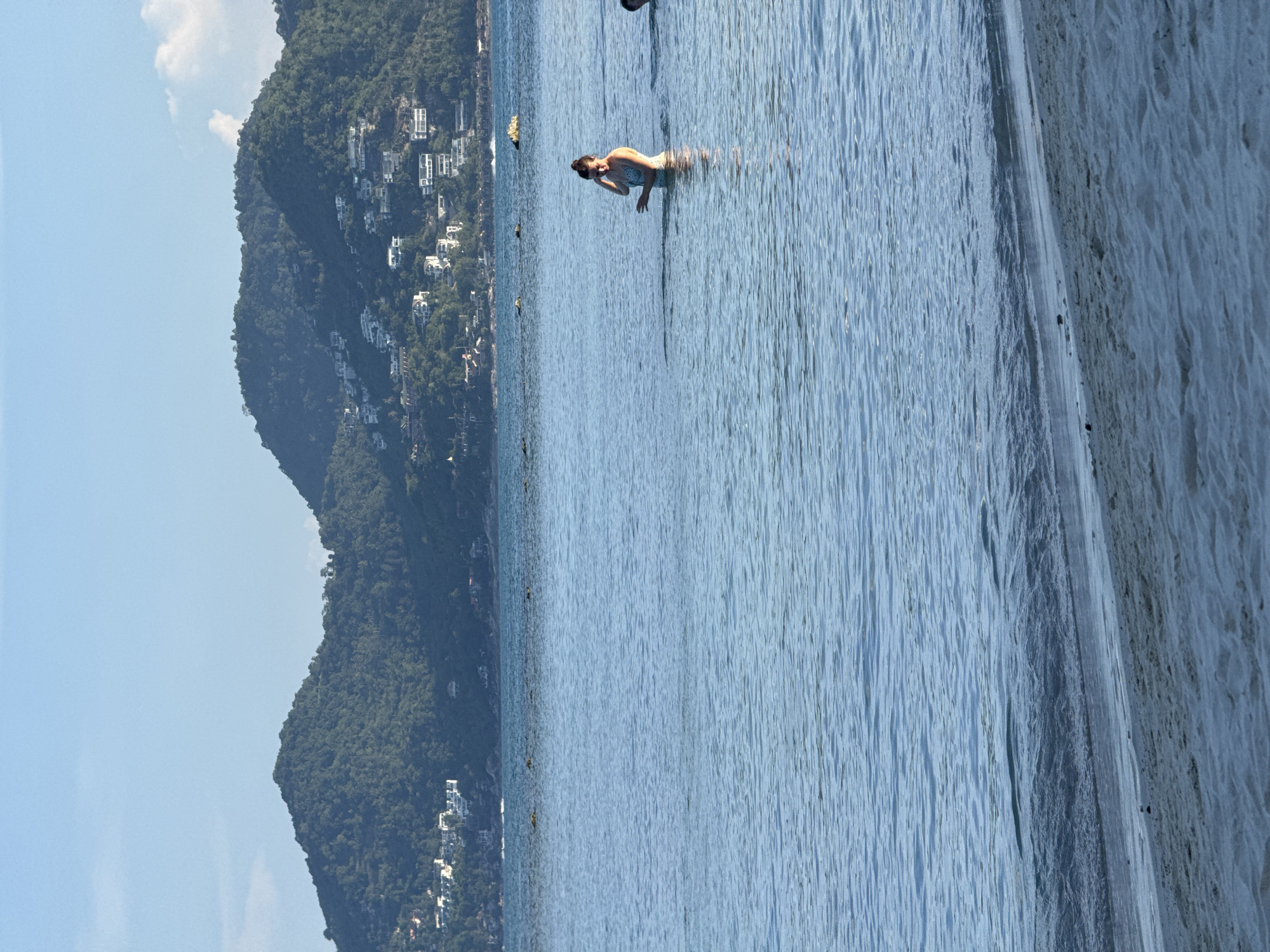A woman stands in the shallow ocean water, facing right. She is wearing a light-colored swimsuit and has her hair pulled back. Her right arm is bent at the elbow, and her hand is raised to her head. The water is calm and blue, with small ripples. In the background, lush green hills covered with houses and trees rise from the sea. The sky above is a pale blue with a few wispy clouds. The foreground shows a sandy beach.