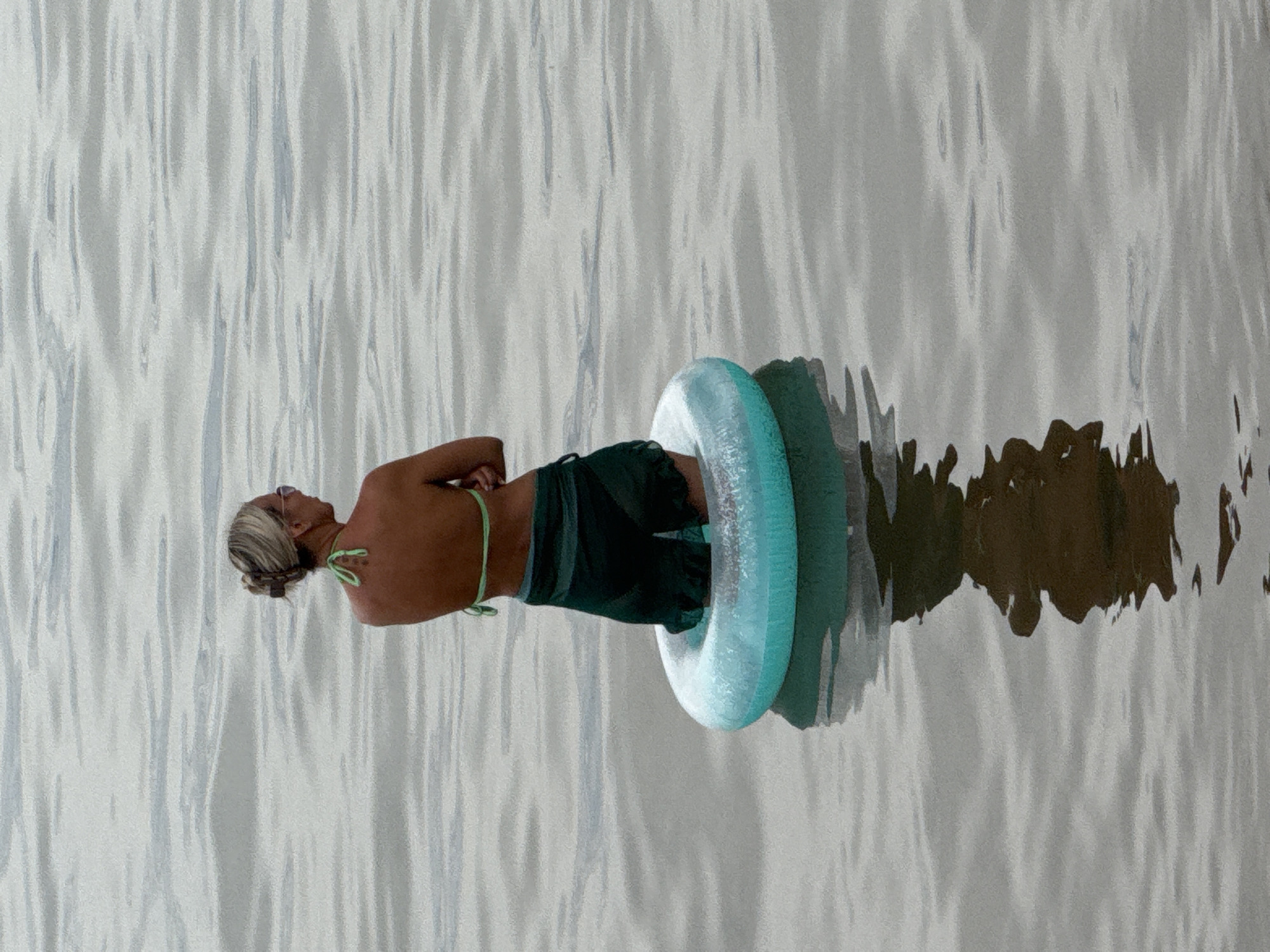 A person with blonde hair pulled back in a bun, wearing sunglasses, a green bikini top, and dark swim shorts, stands in the water of Lake Kulunda in Yarovoye, Russia. They are seated within a light blue inflatable ring. The person is facing away from the camera, looking out at the water. The water is calm and reflects the overcast sky. The scene appears to be a peaceful moment of relaxation.