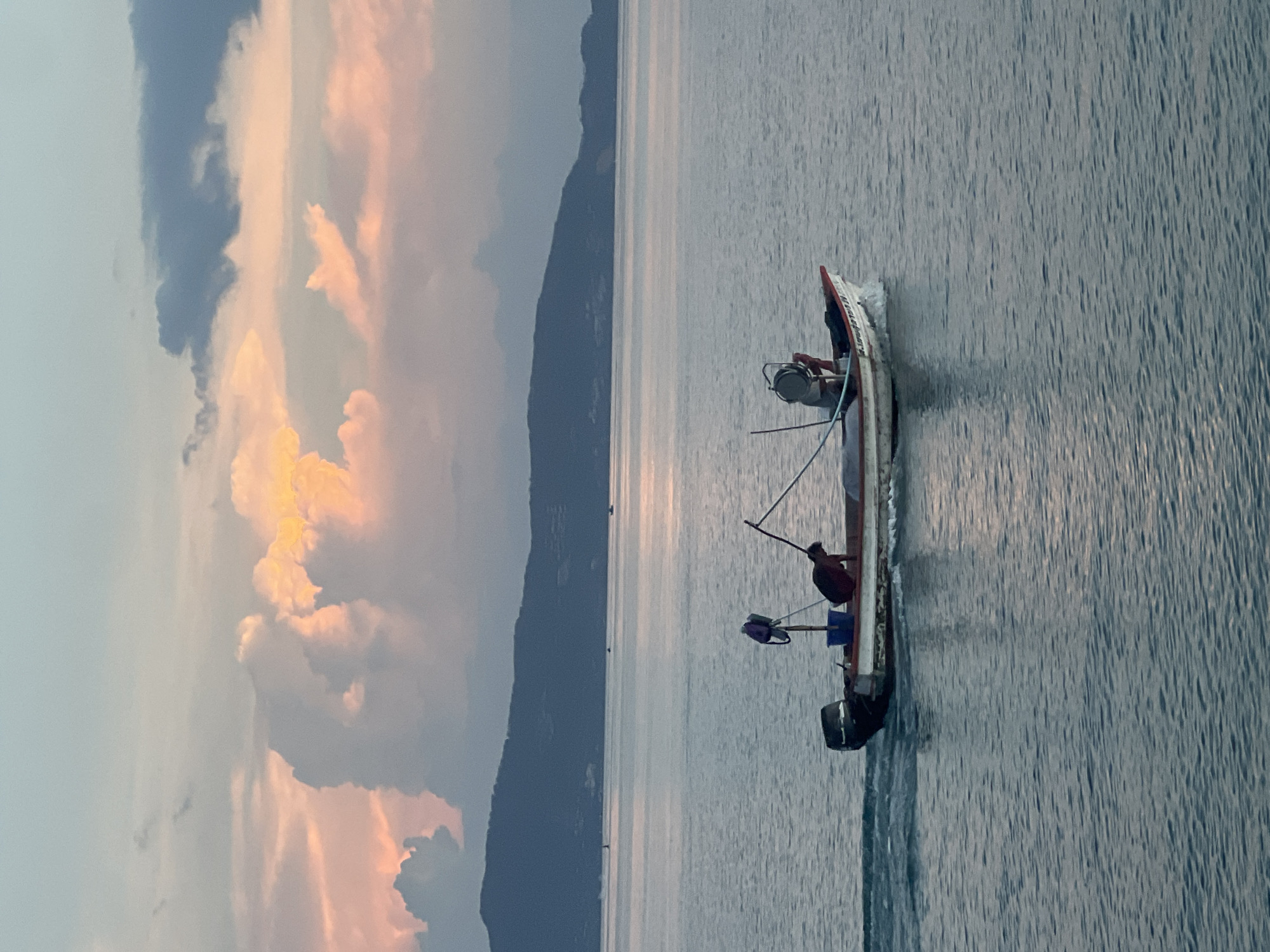 The image shows a small fishing boat with two individuals on board, moving across a calm body of water. The boat is white with red trim and has an outboard motor at the stern. One person, wearing a dark shirt, is seated in the boat, while another person, wearing a red shirt, is leaning over the side. A blue bucket and a purple object are also visible in the boat. The water is a muted blue-gray, reflecting the soft light of the sky, which is filled with large, dramatic clouds in hues of pink, orange, and gray. In the background, a silhouetted mountain range with some visible structures suggests a coastal or island landscape, characteristic of Ko Pha-ngan District, Thailand. The overall scene captures a serene moment during either sunrise or sunset.