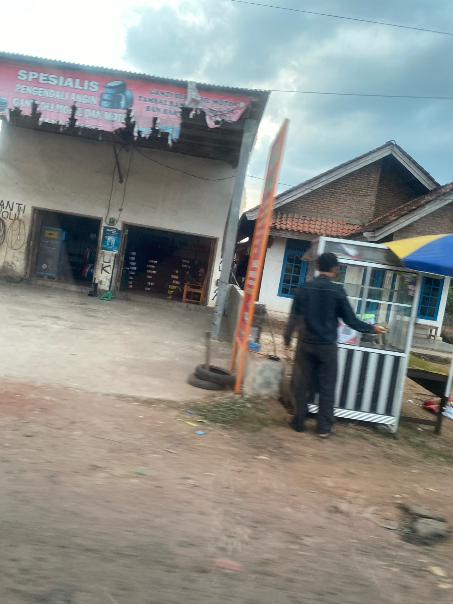 The image shows a street scene in Toto Katon, Indonesia, with a small business establishment on the left and a residential structure on the right. The business, identified by a torn red and white banner reading "SPESIALIS PENGENDALI ANGIN GANTI OLI MOTOR DAN MOTOR", appears to be a motorcycle repair shop and tire service. There are two open doorways leading into the shop, with tires visible inside. To the right of the shop, a man in a dark long-sleeved shirt and dark pants is standing by a food cart with black and white vertical stripes. The food cart has a blue and yellow umbrella over it. A house with a terracotta tiled roof and blue-framed windows is visible behind the food cart. The ground in front of the establishments is unpaved and appears to be a dirt road. Several tires are stacked on the ground in front of the shop. The sky is overcast.