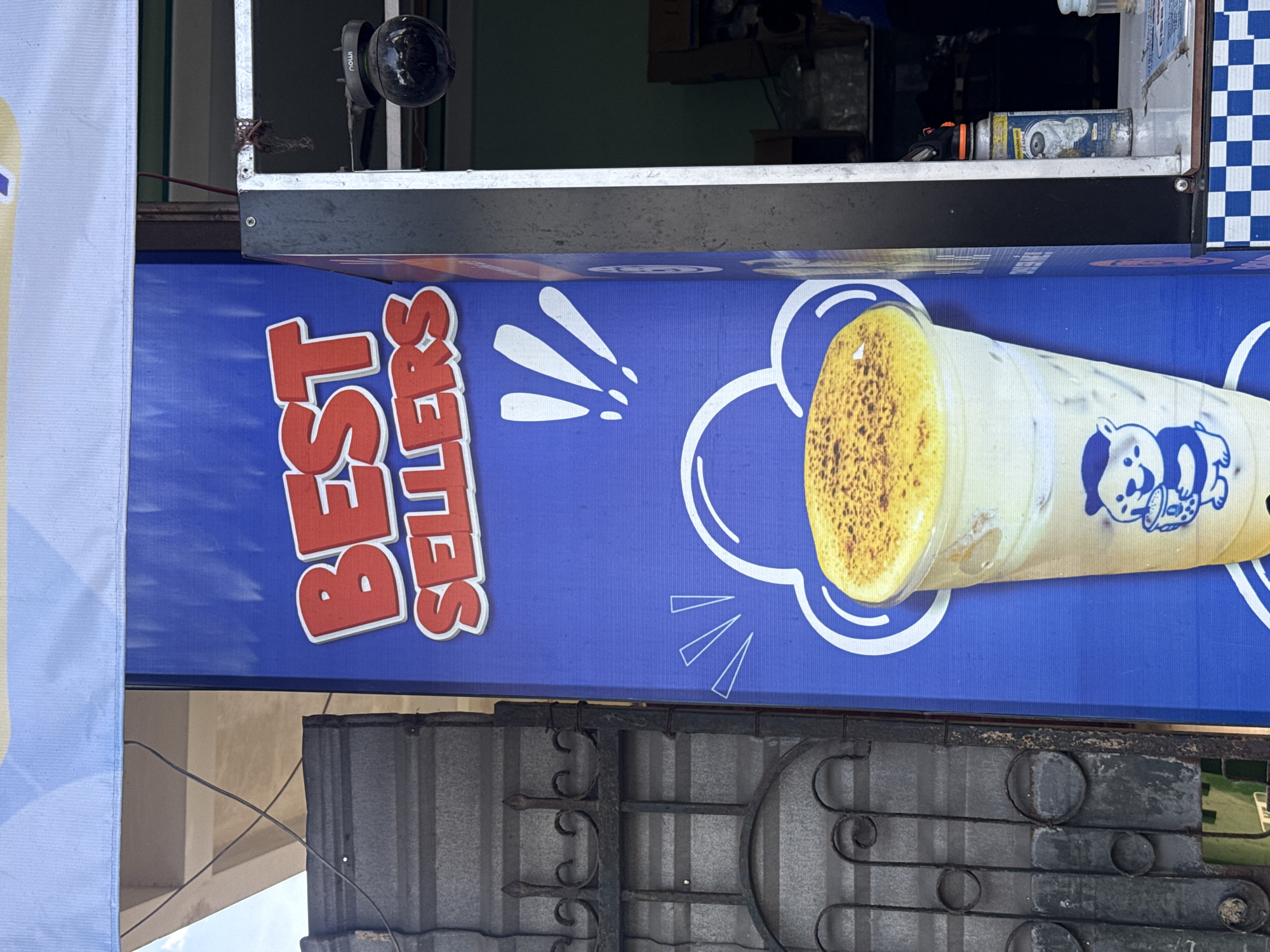 A large, blue banner advertising "BEST SELLERS" in red, bubbly lettering is displayed on the exterior of a building. The banner features an illustration of a large, clear plastic cup filled with a creamy beverage topped with a golden powder, and a cartoon bear character holding a smaller cup. To the left of the banner is a dark, ornate metal gate. To the right, a portion of a kiosk is visible, with a security camera mounted above it. The scene appears to be in Nha Trang, Vietnam, given the presence of a can with Vietnamese labeling and the overall architectural style. The lighting suggests it is daytime.
