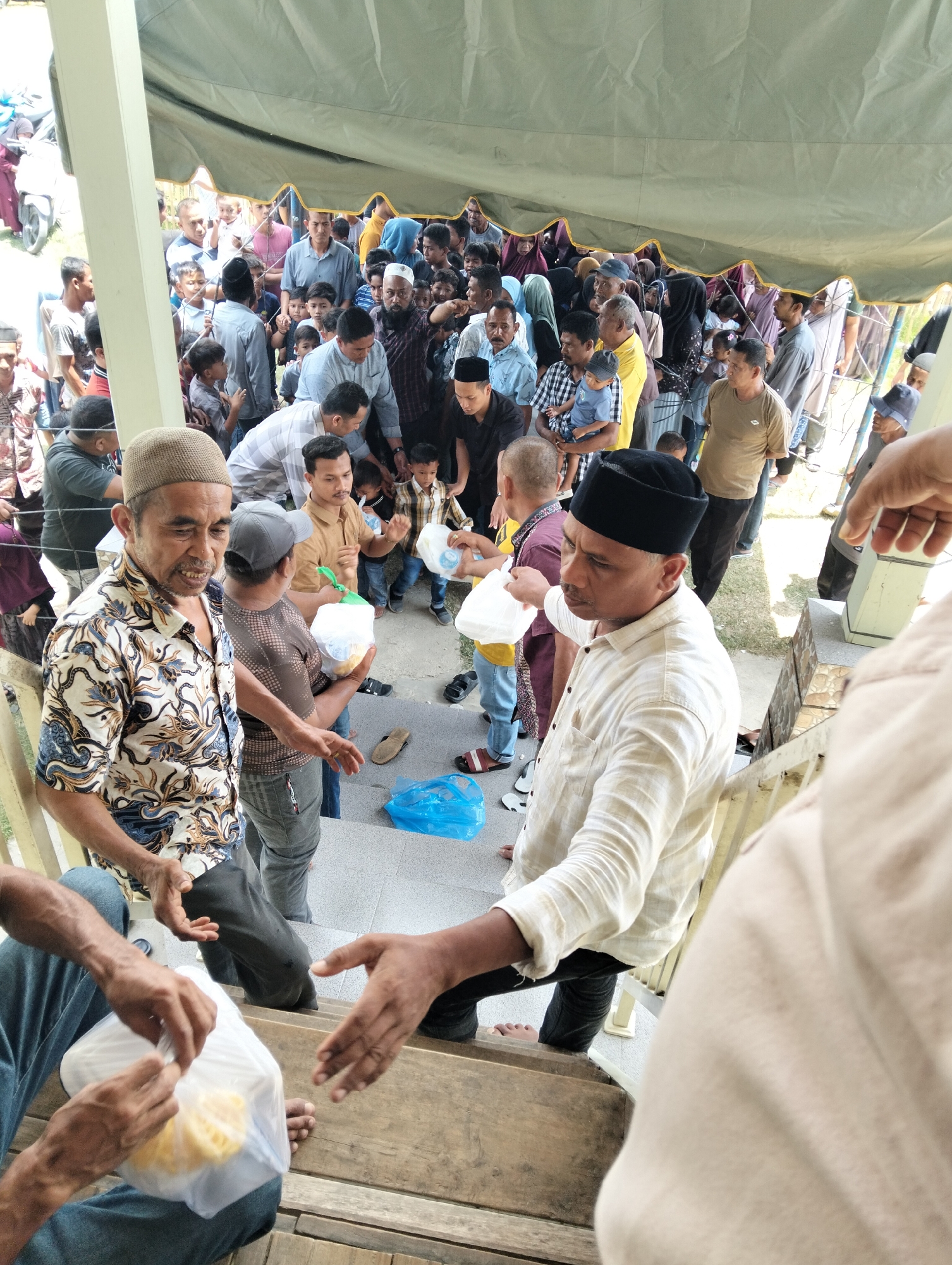 The image depicts a gathering of people, likely receiving aid, on a set of wooden stairs. The scene appears to be outdoors, under a large tent with a light green fabric. The people are a mix of men, women, and children, many dressed in casual attire, with some women wearing headscarves. Several individuals are holding white plastic bags, some containing yellow items, suggesting they are receiving food or goods.
In the foreground, two men are prominently featured. The man on the left is wearing a patterned shirt and a light brown cap, and he is reaching out his hand. The man on the right, wearing a light-colored collared shirt and a black cap, is also extending his hand, possibly in a gesture of giving or receiving. The interaction between them, as well as with others in the vicinity, suggests a distribution or exchange is taking place.
The background is filled with a crowd of people, indicating a significant number of attendees. Some are looking towards the foreground, while others are engaged in conversation or looking in different directions. The environment suggests a community event or charitable distribution. The presence of a light blue plastic bag on the stairs and a sandal on the ground suggests a somewhat informal setting. The context provided suggests this event is taking place in the city of Grong-Grong, Indonesia.