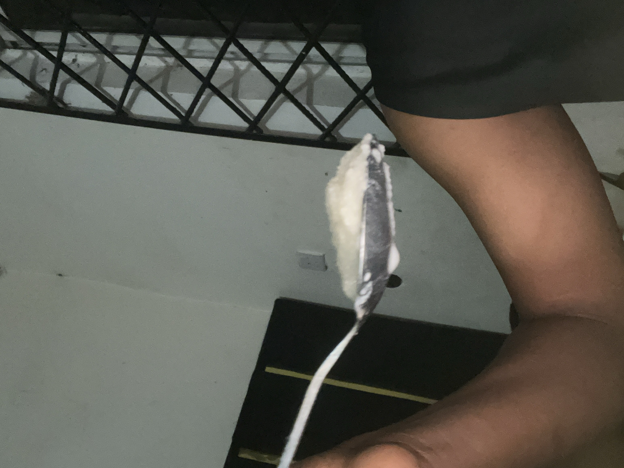 A person is holding a white charger. The charger's head is covered in a white, foamy substance. A security bar, a wall with a power outlet, and a black door with a yellow horizontal stripe are visible in the background. The image's location is unknown; there is no reliable information to place it in Lokoja, Nigeria.