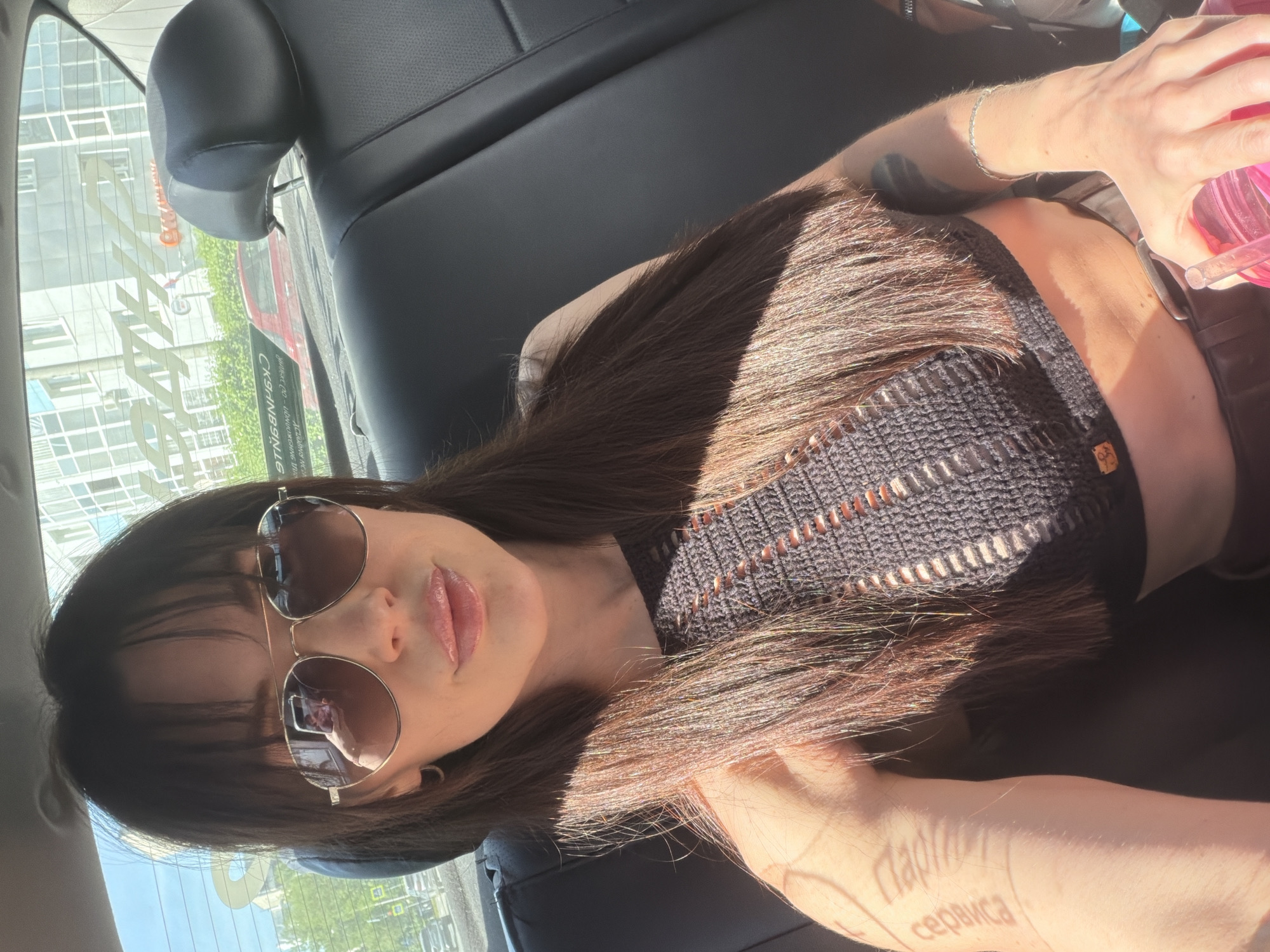 The image depicts a woman wearing sunglasses and a black crocheted top, sitting in the back of a car. She is holding a pink drinking container. Through the car window, the building across the street displays signage in Russian, including the word "Пермь" (Perm), indicating the location as the city of Perm, Russia. The sign displays "CКЯНИНГ CЕРВИСА". The woman's arm has a tattoo displaying Russian Cyrillic script.