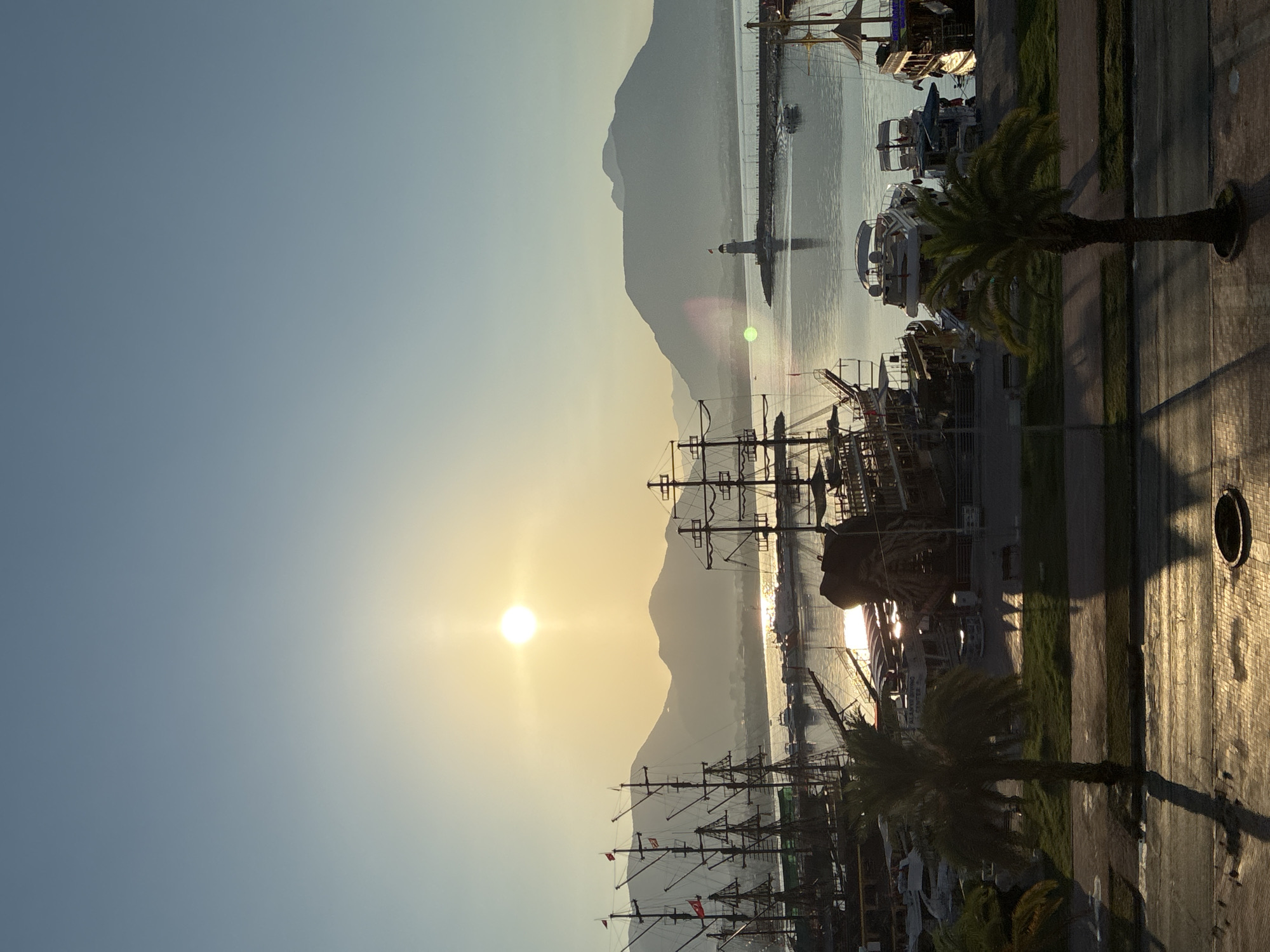 The image depicts a scenic view of Alanya, Turkey, at sunrise or sunset. In the foreground, a paved area with shadows of palm trees suggests a waterfront promenade. Two palm trees are visible, with their fronds casting long shadows. Beyond this, a grassy area separates the promenade from a harbor.
The harbor is filled with various boats, prominently featuring several large, wooden sailing ships with multiple masts, reminiscent of pirate ships or historical vessels. Alongside these, modern motorboats and yachts are docked. The water reflects the warm glow of the sun.
In the mid-ground, a lighthouse stands on a pier extending into the sea, guiding ships. The background is dominated by a range of mountains, silhouetted against the bright sky. The sun, a brilliant orb, is low in the sky, casting a warm, golden light over the entire scene. The sky transitions from a soft yellow near the horizon to a pale blue at the top of the frame. The overall impression is one of tranquility and natural beauty, characteristic of a coastal city like Alanya.