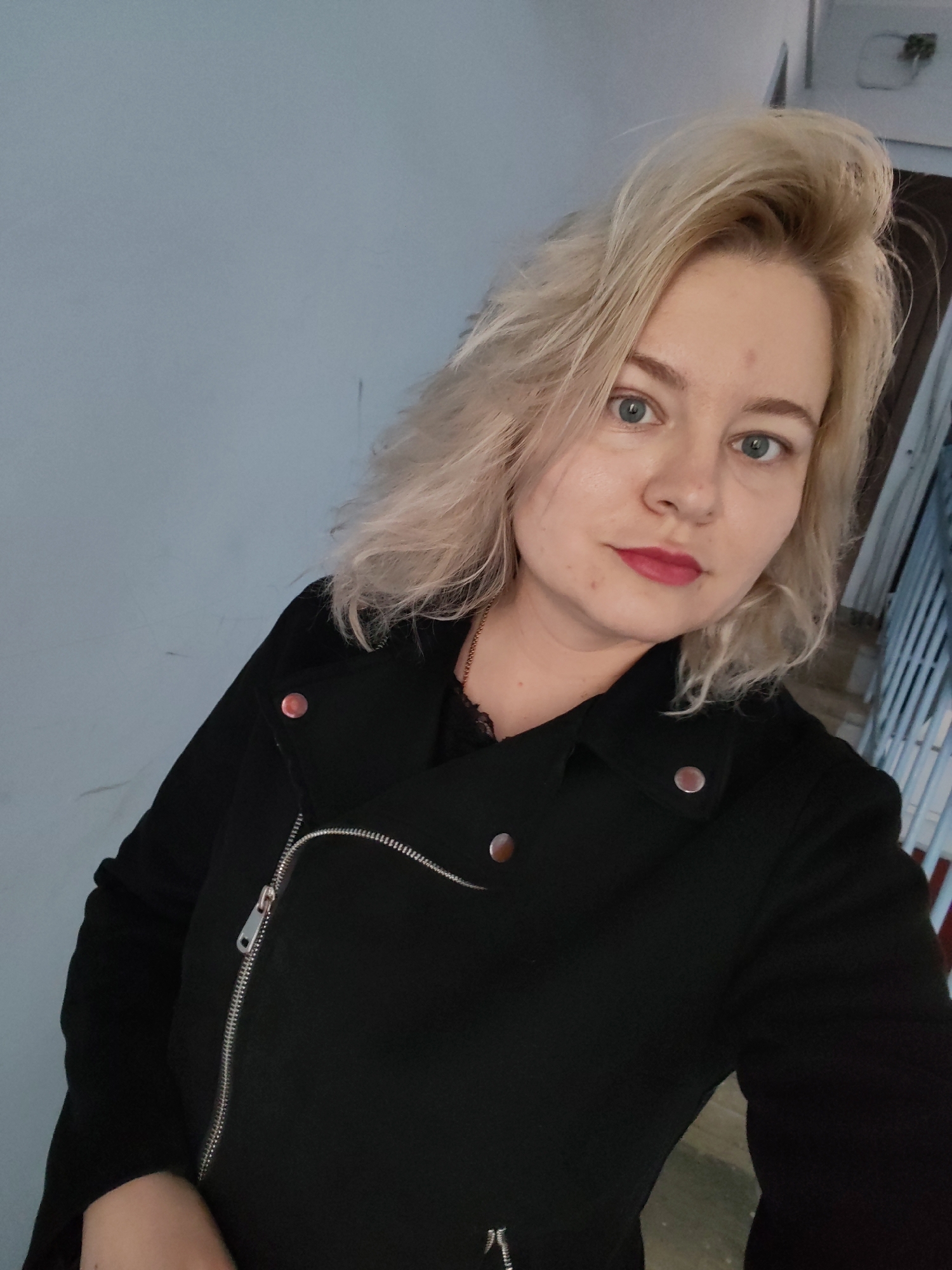 A fair-skinned adult female, appearing to be in her 20s or 30s, is captured in a close-up, chest-up selfie, looking directly into the camera with a neutral expression. She has shoulder-length, wavy, platinum blonde hair styled with volume. Her eyes are blue, and she is wearing red lipstick; minor blemishes are visible on her forehead and left cheek. She is dressed in a black moto-style jacket featuring an asymmetrical silver zipper running diagonally across the chest, and multiple silver-toned snap buttons on the lapels and shoulders. A dark, possibly black, lace-trimmed garment and a thin gold-colored chain necklace are visible beneath the jacket's opening. The background consists of a plain light grey-blue wall. To the right of the subject, a dark brown door frame and a section of a light-colored metal railing, consistent with an indoor hallway or stairwell, are partially visible. The specific city of Izhevsk, Russia, cannot be visually confirmed from the image.