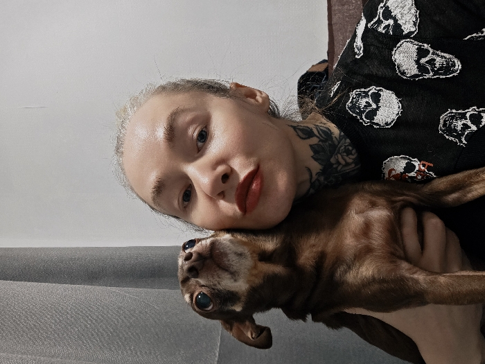 A woman with pale skin, blonde hair, and a flower tattoo on her neck is holding a brown chihuahua. The woman is wearing red lipstick and a black shirt with white skull designs. She is holding the dog close to her chest, with her head resting against the dog's head. The dog is looking up at the camera with wide eyes. The background is a plain white wall with a gray couch in the lower left corner. The image is rotated to the left.