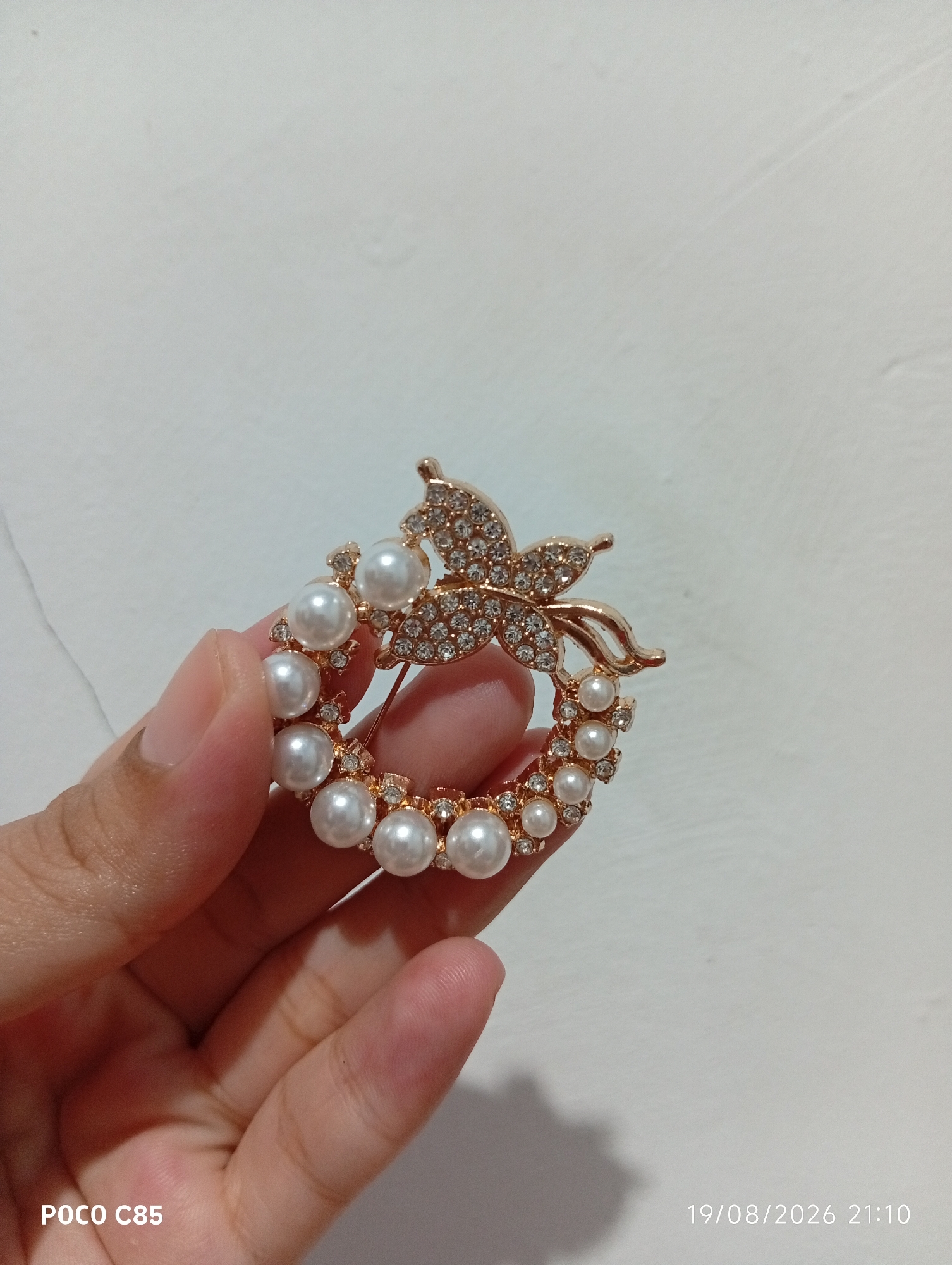 A close-up shot shows a hand holding a gold-toned brooch adorned with pearls and rhinestones in the shape of a wreath with a butterfly at the top. The brooch is circular and features various sizes of pearls and small, clear rhinestones. The hand is gently holding the brooch from the bottom left, with the fingers partially obscuring the lower portion of the accessory. The background is a plain, off-white textured surface, possibly a wall. In the lower left corner of the image, the text "POCO C85" is visible, and in the lower right corner, a date and time stamp reads "19/08/2026 21:10". The image quality suggests it was taken with a mobile phone camera. The location is specified as South Tangerang, Indonesia.