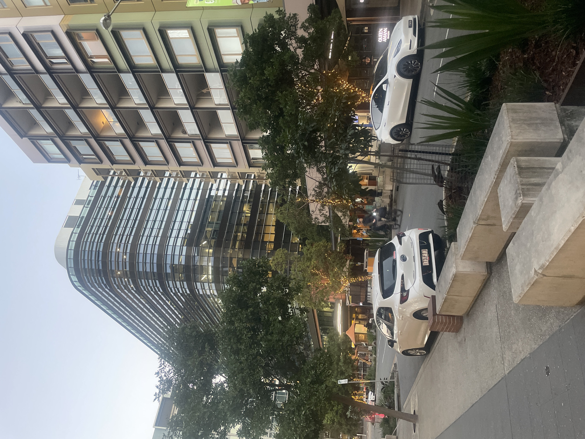 A street scene in Brisbane, Australia, captures modern architecture and urban life at dusk. The foreground features a paved sidewalk with concrete blocks and planters containing green foliage. A white Toyota Corolla hatchback is parked on the street, facing away from the camera, with its license plate clearly visible. Next to it, a white Tesla Model Y is parked, also facing away. A black bicycle rider is visible in the background on the street.
Dominating the background are two prominent buildings. To the left, a contemporary office building with a curved facade and numerous horizontal windows reflects the soft evening light. To the right, a multi-story apartment building displays a grid of balconies with glass railings and a mix of lit and unlit windows. The street is lined with trees, some adorned with decorative string lights, hinting at an evening ambiance. The sky above is a clear, pale blue, indicative of twilight. The overall impression is one of an urban environment transitioning from day to night.