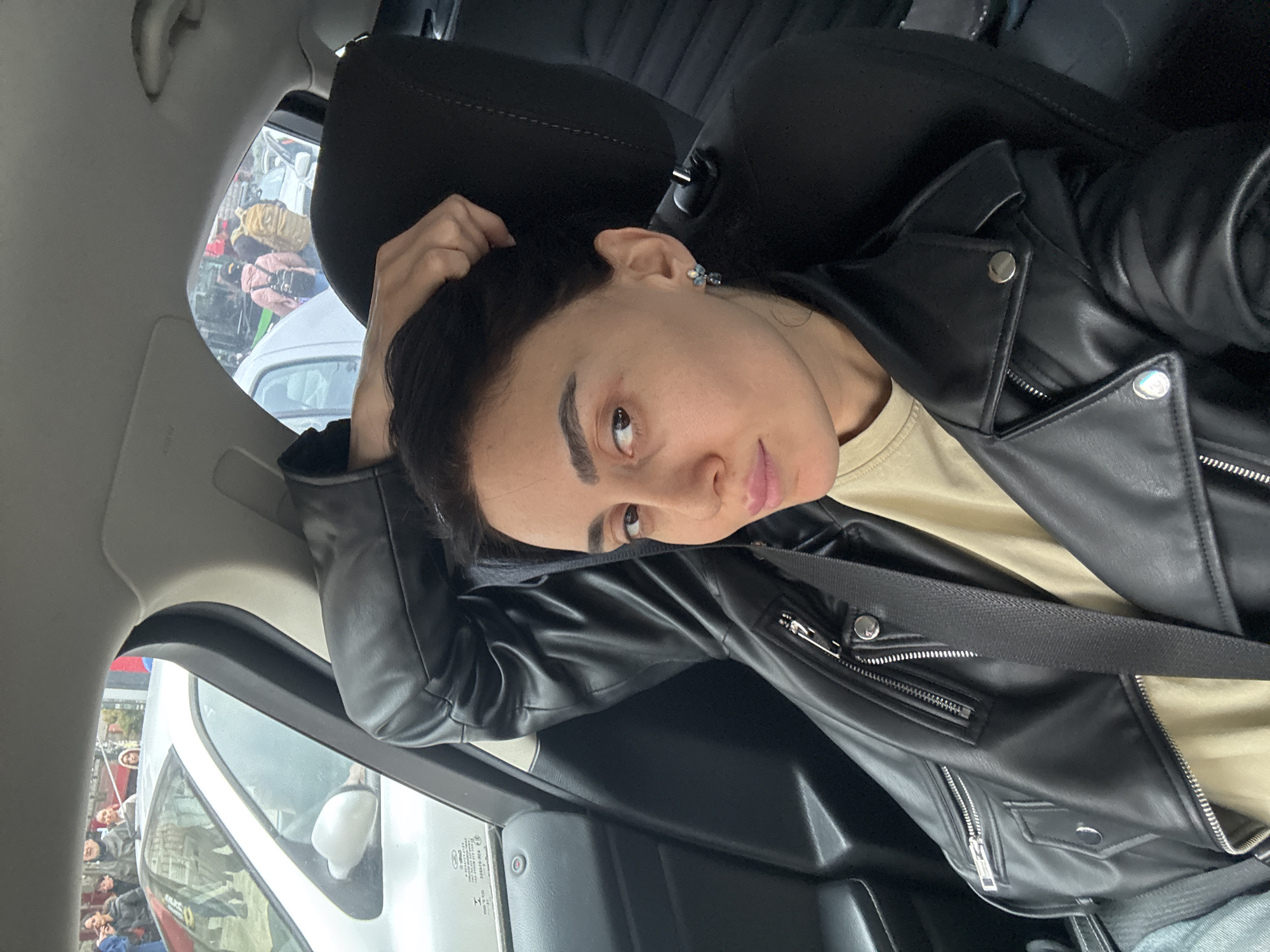 A female individual is depicted in a close-up, front-facing selfie taken from inside the front passenger seat of a vehicle in Tbilisi, Georgia. She has dark hair, dark eyebrows, and brown eyes, looking directly at the camera with a subtle expression. Her right hand is positioned at the back of her head, with fingers touching her hair. She wears a black leather jacket with multiple silver zippers and snaps over a light beige t-shirt, secured by a black seatbelt across her chest. A multi-stone earring, appearing blue and silver, is visible in her right ear. The car's interior is dark-toned, featuring black seating and door panel elements. Through the vehicle's windows, an urban street scene is visible, including a silver sedan positioned alongside, and numerous pedestrians on a sidewalk. Individuals in the background are wearing jackets, and some are carrying backpacks, indicating an active outdoor environment.