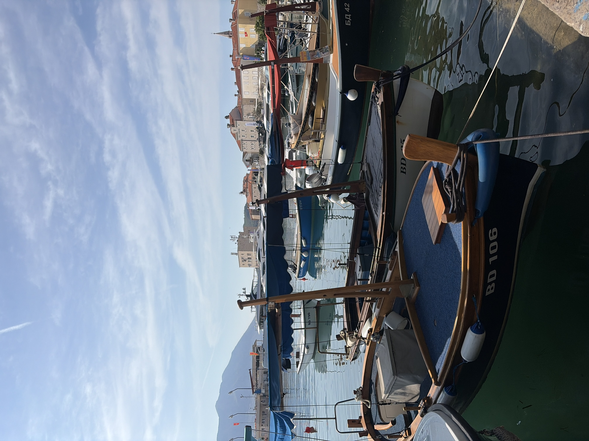 In Budva, Montenegro, a waterfront scene unfolds under a sky dotted with wispy clouds and a faint contrail. The foreground is dominated by several docked wooden boats, their hulls painted in varying shades of dark blue and white, some featuring canvas awnings for shade. One boat in the immediate foreground displays the registration "BD 106" in white lettering. Mooring ropes and blue fenders are visible.
In the middle ground, more boats of similar design are moored in a calm, reflective body of water. A few larger yachts with sleek white hulls are visible further out in the harbor.
The background reveals the distinctive architecture of Budva's old town, characterized by buildings with red-tiled roofs and stone facades. A prominent tower with a cross on top, likely part of a church, stands out against the cityscape. In the distance, a hazy mountain range provides a natural backdrop. The overall impression is one of a peaceful harbor on a sunny day. There are no people clearly visible in the image.