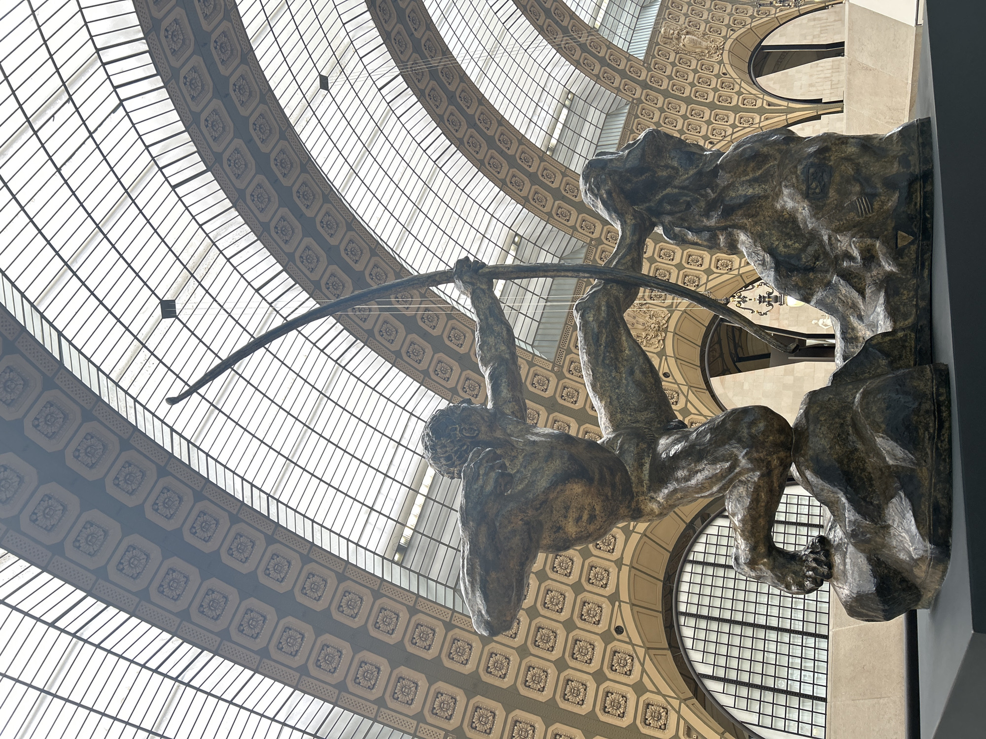 The photograph, taken from a low angle, depicts a bronze sculpture of a male figure drawing a bow and arrow, set against the backdrop of a glass-paneled dome and intricately patterned ceiling. The figure, weathered and textured, is positioned with the bow arching toward the top left corner of the frame. The glass dome features a grid-like pattern with numerous individual panes. The surrounding ceiling area is adorned with a pattern of repeating square motifs, each containing a stylized floral design. Behind the sculpture and to the right, the ceiling design continues with occasional arch-shaped cutouts. The setting evokes the grandeur of a classical building interior, strongly suggesting that the location is the Musée d'Orsay in Paris, France, known for its Belle Époque architecture and art collection.